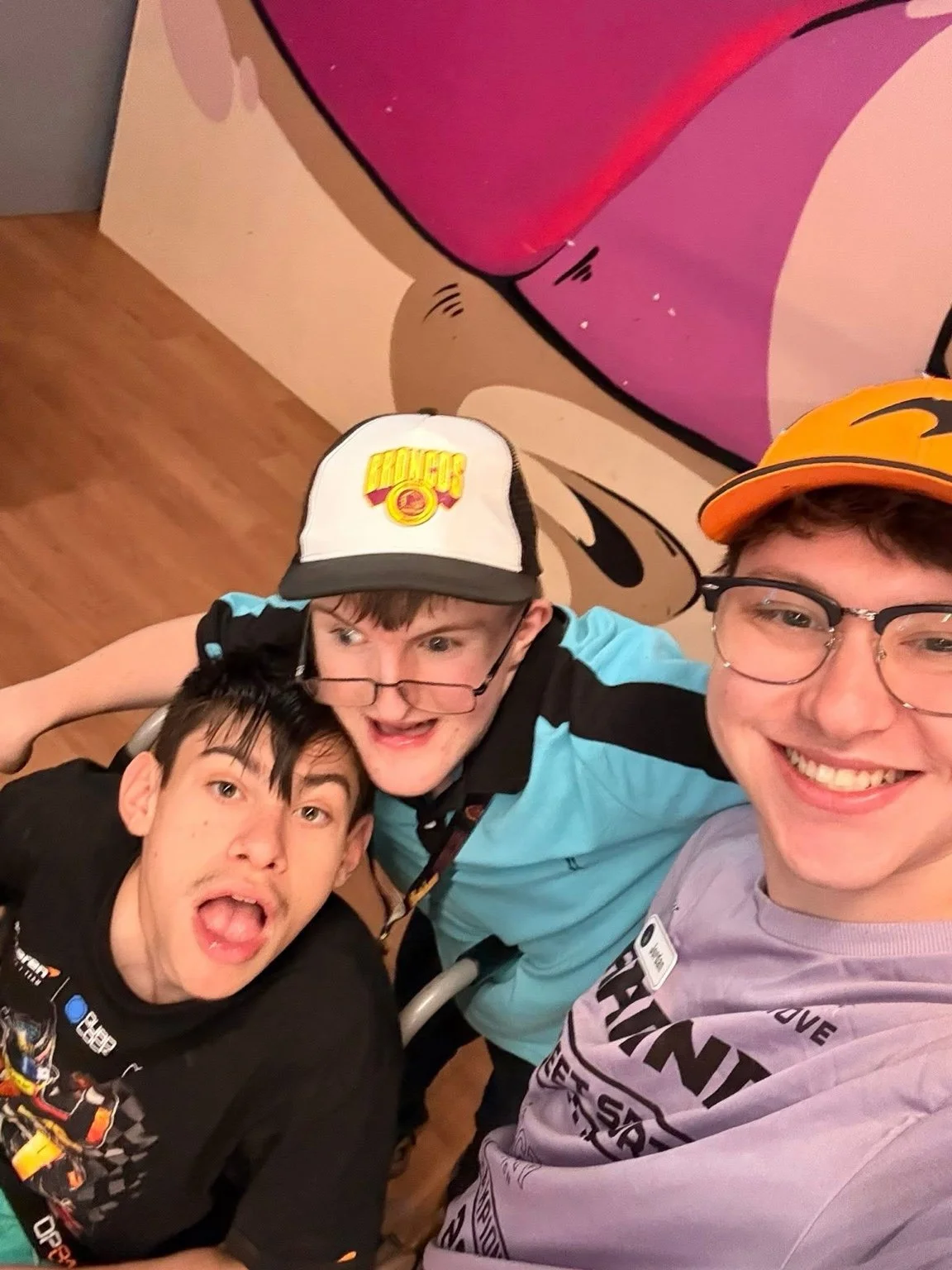 Three young men taking a selfie together, smiling and making playful faces, with a colorful cartoon wall in the background.