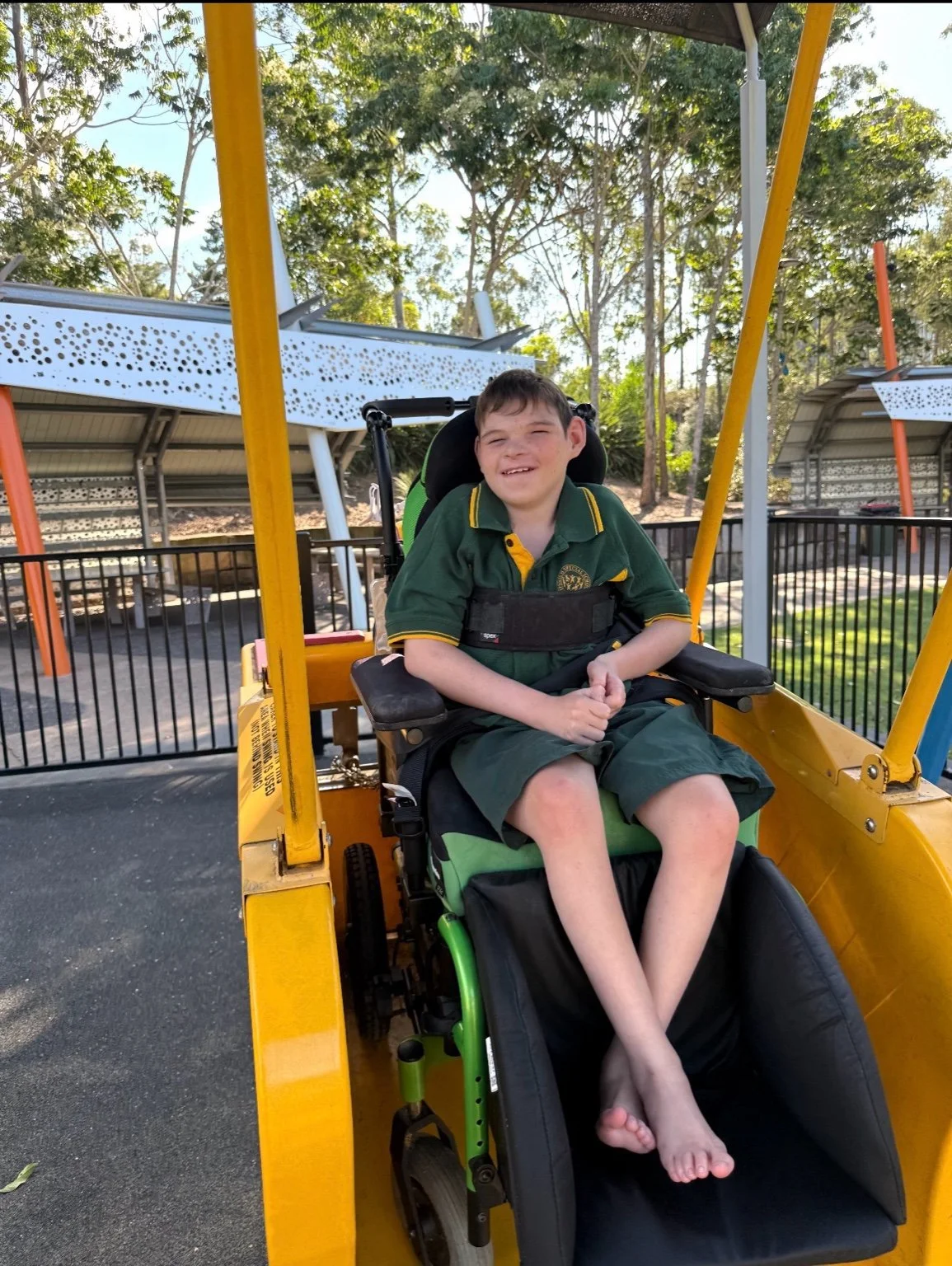 A young boy in a wheelchair smiling and sitting on a yellow ride at an outdoor playground with metal structures and green trees in the background.