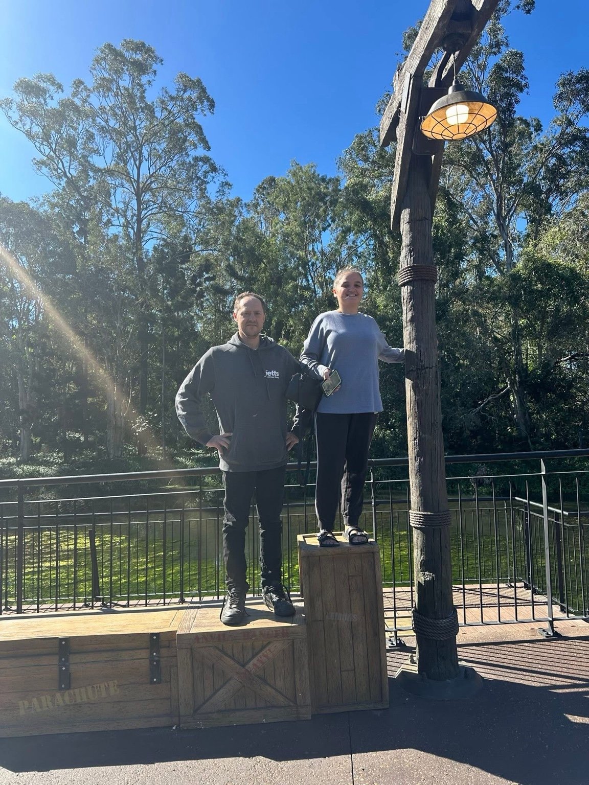 A man and woman standing on a wooden podium outdoors, with a forest and blue sky in the background. The woman is standing on a smaller platform, smiling, holding a phone, and the man stands on a lower platform with his hands on his hips. There is a l