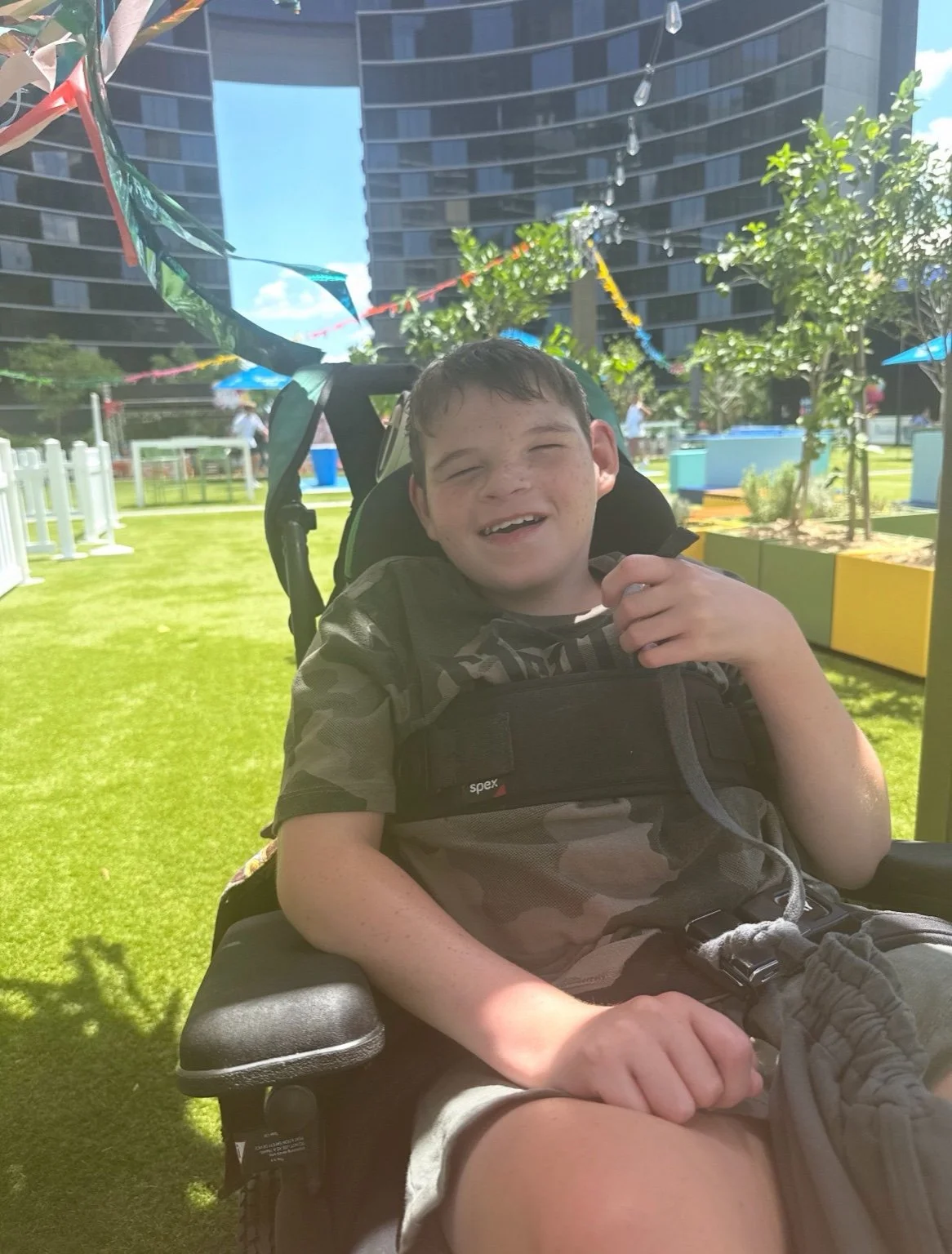 A smiling boy with freckles in a wheelchair outdoors on a sunny day, with colorful banners and a modern urban background.