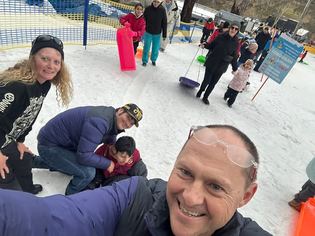 Group of people at an outdoor ice skating rink, some preparing to skate or rest, and taking a selfie with the ice rink in the background.