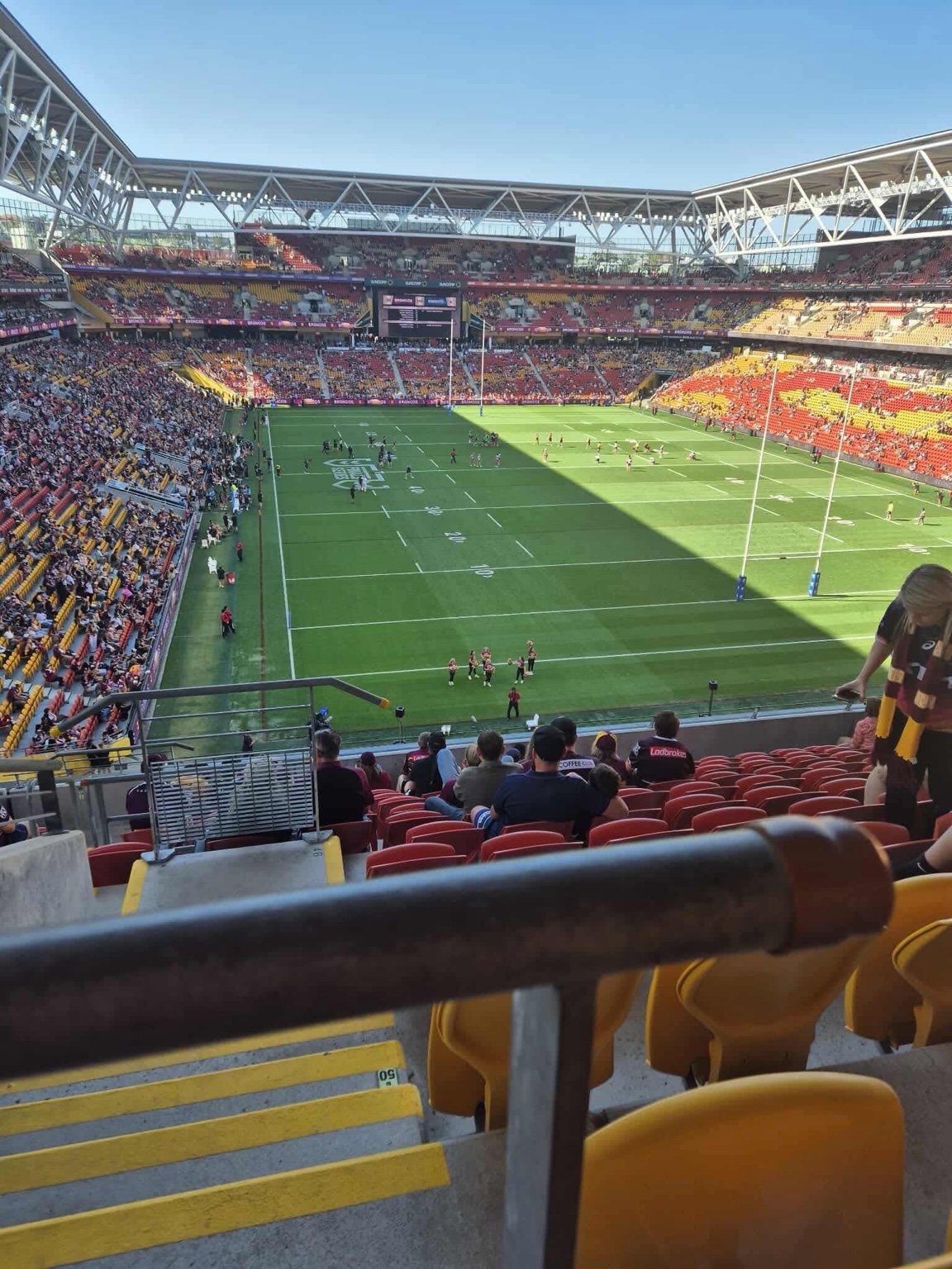 Inside a stadium, view of a football field with players and spectators, with a modern roof structure and bright sunlight casting shadows.