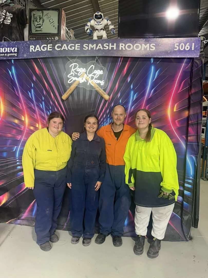 Four people standing in front of a colorful backdrop for Rage Cage Smash Rooms, with a sign above reading "Rage Cage Smash Rooms." Two of the individuals are wearing yellow and black uniforms. The person in the middle is wearing an orange shirt, and 