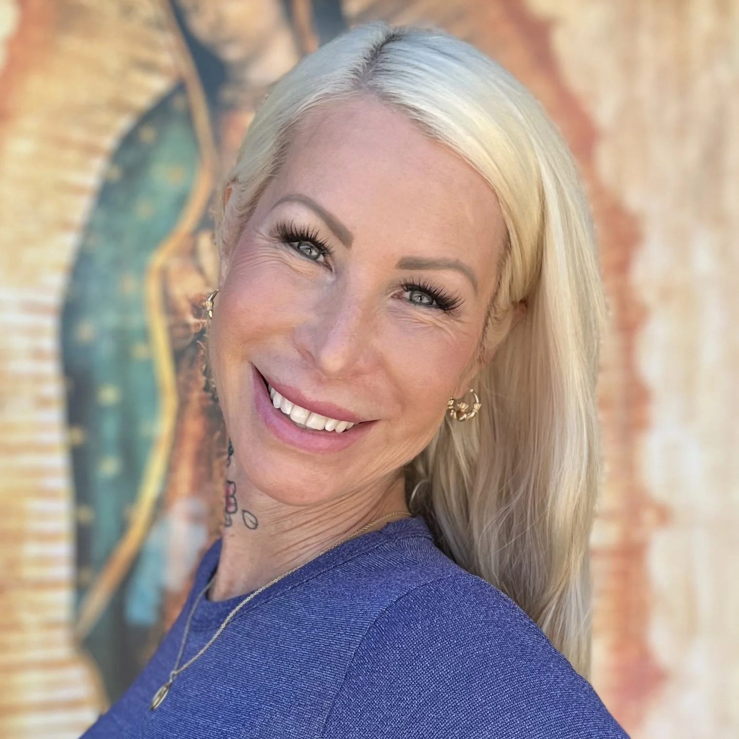 A smiling woman with long blonde hair, wearing a blue top and gold earrings, standing in front of colorful abstract artwork.