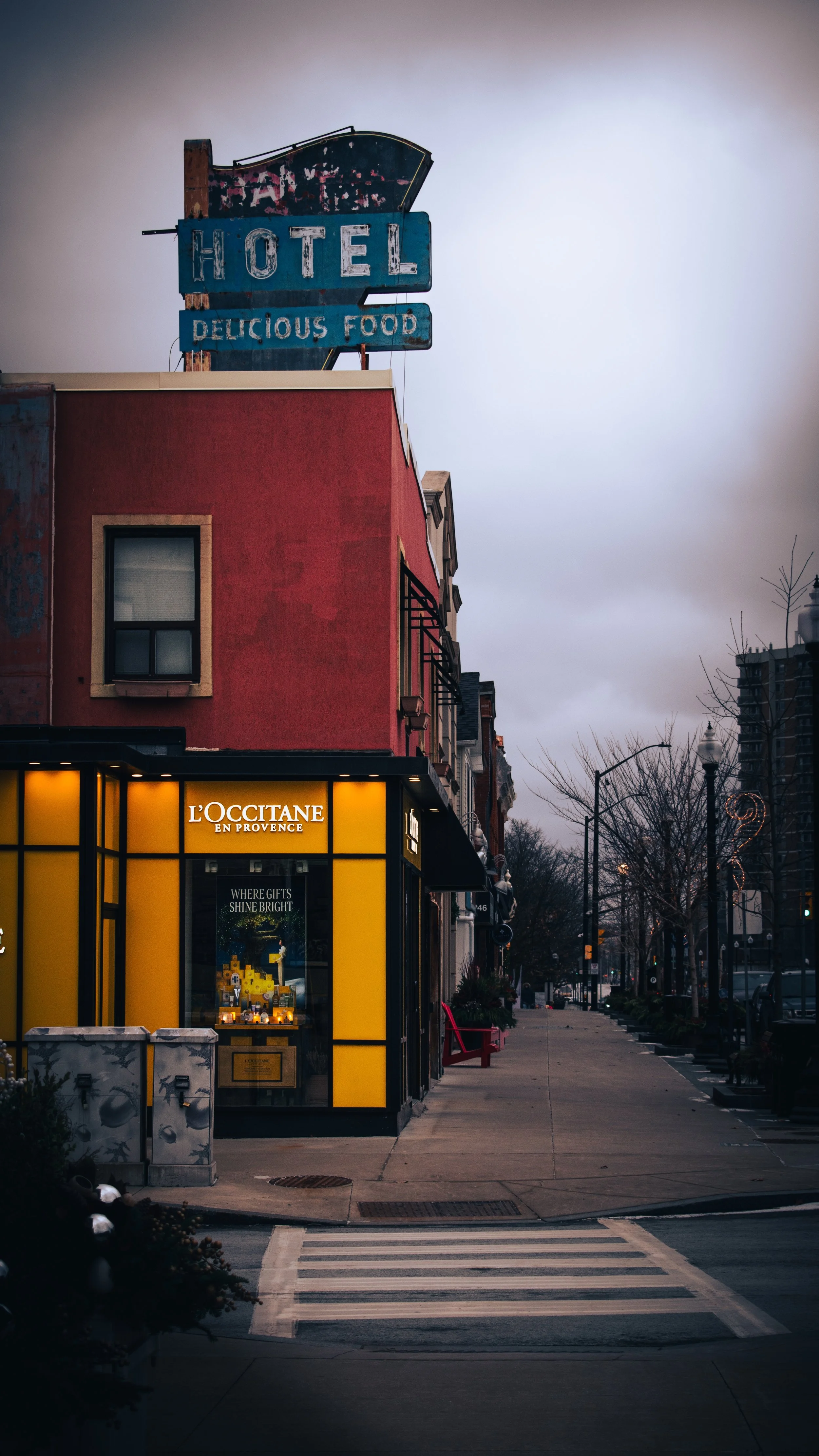 Old neon hotel sign with faded lettering, a modern storefront with yellow and black signage for L'Occitane en Provence, and a city sidewalk with bare trees and lampposts on a cloudy day.
