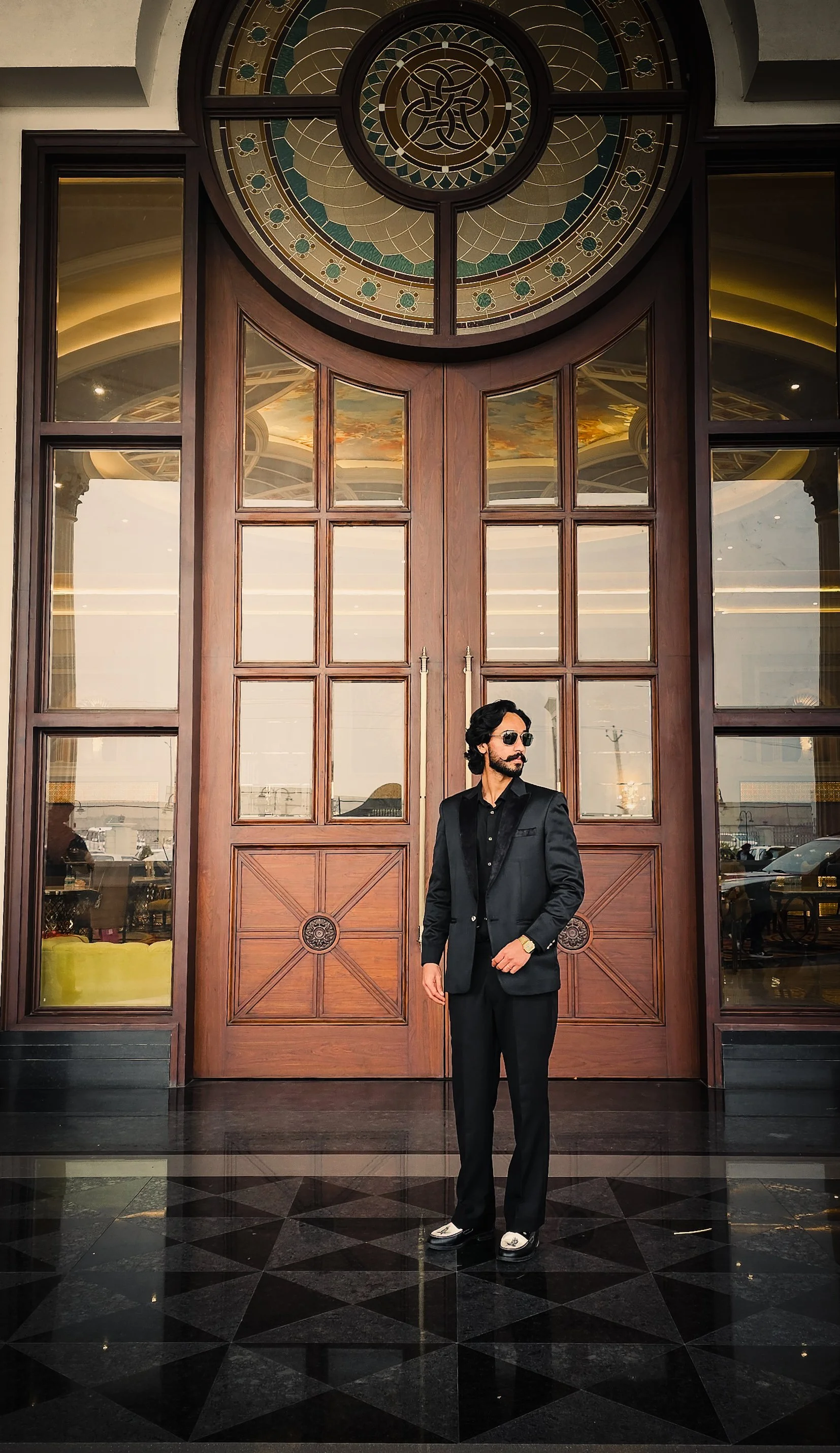 A man in a black suit, black shirt, and sunglasses standing in front of large wooden double doors with glass panels and an intricate stained-glass semi-circular window above, reflecting a stylish interior and an outdoor scene.