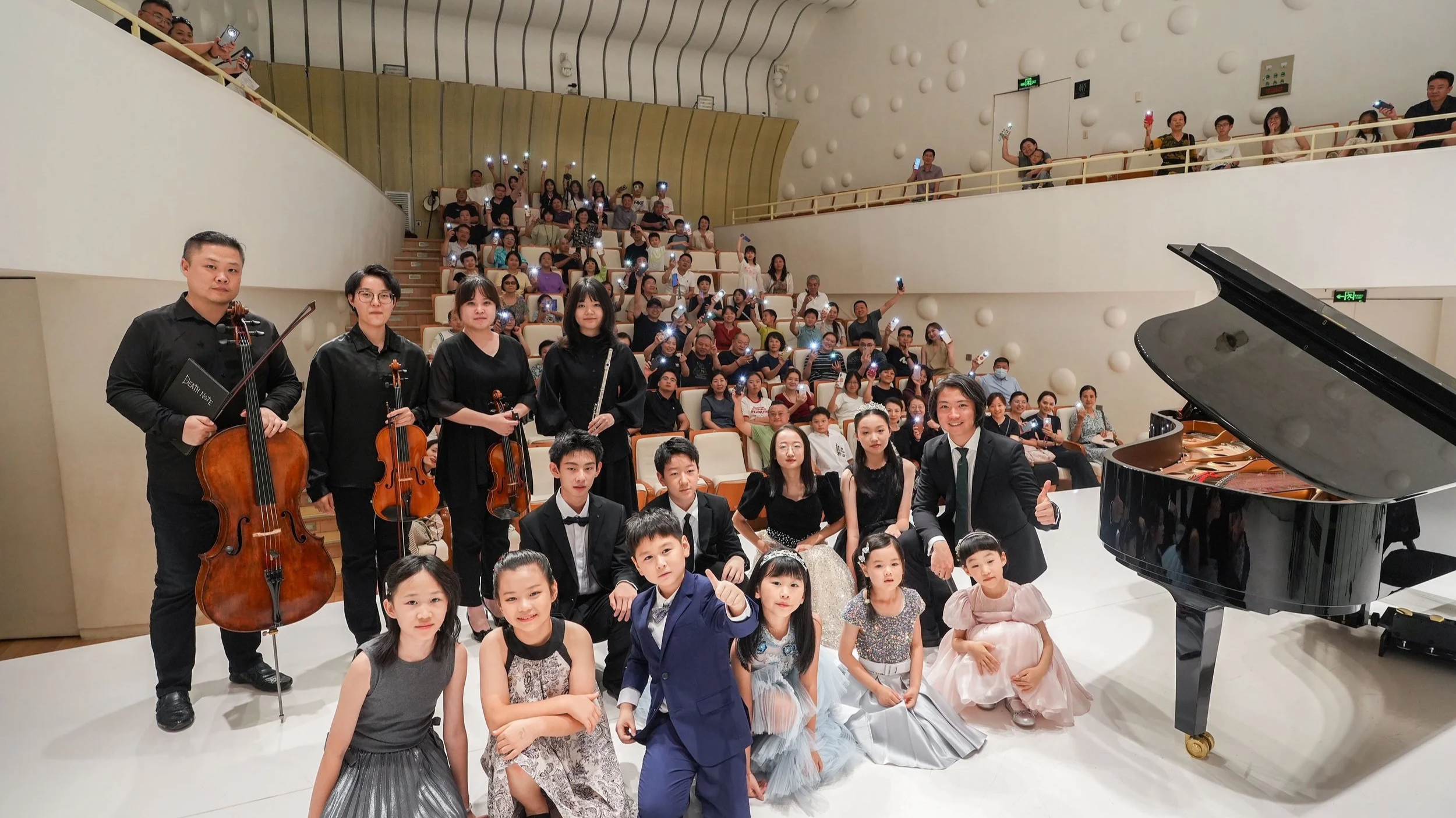 Group photo of young musicians and conductor on stage after a performance with an audience in the background.