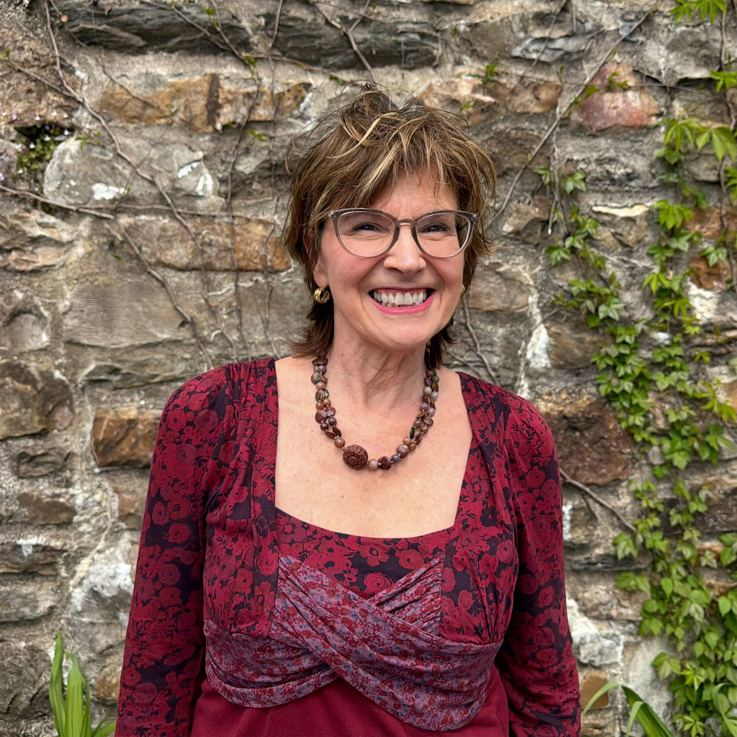 A smiling woman with short brown hair and glasses, wearing a red and black floral dress and a beaded necklace, standing in front of a stone wall with some green ivy growing on it.
