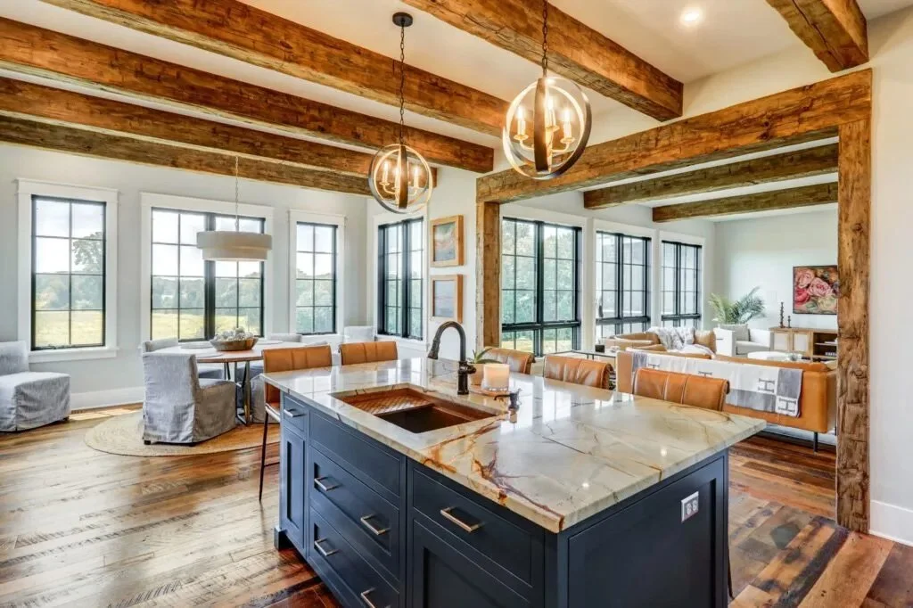 Open-concept living and dining area with exposed wooden beams on the ceiling, large windows allowing natural light, and a kitchen island with a marble countertop.
