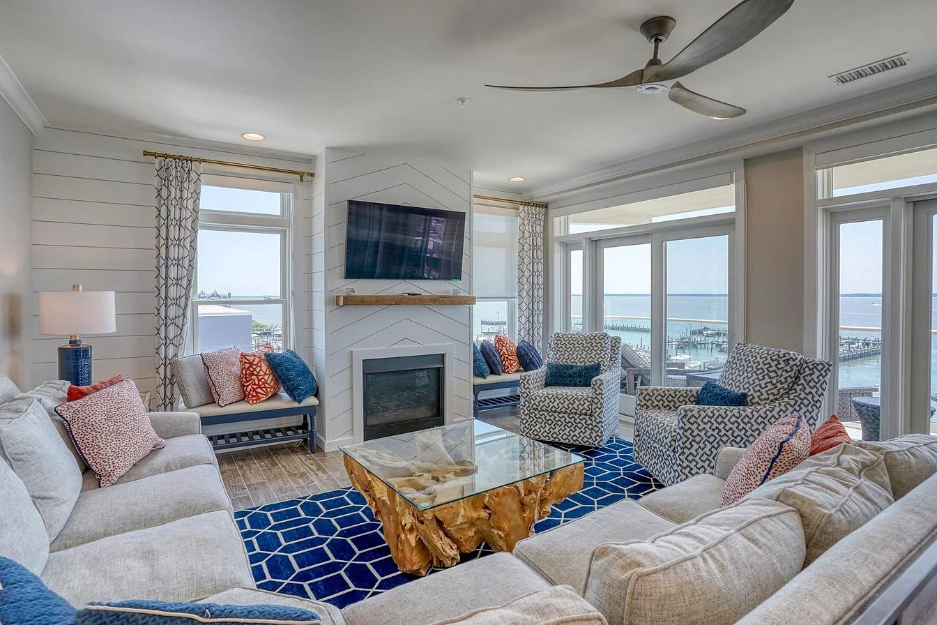 Living room with large windows overlooking a marina, featuring a sectional sofa, patterned armchairs, a glass coffee table with a wooden base, and a television mounted above a fireplace.