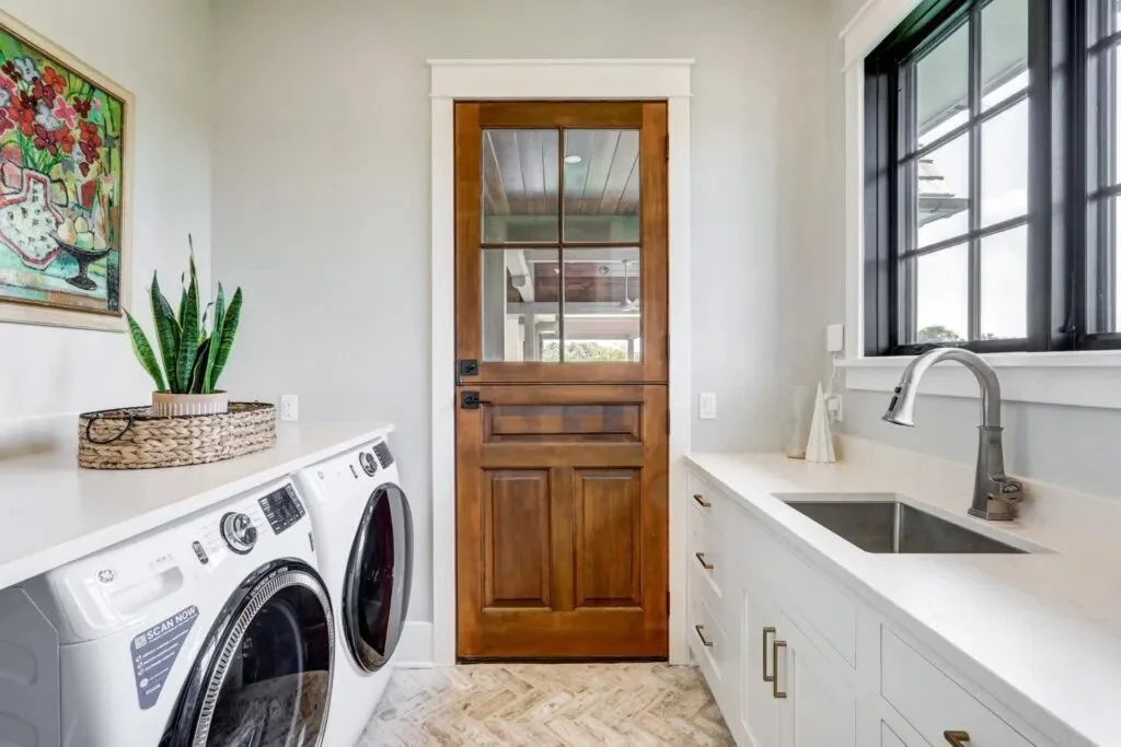A laundry room with white cabinets, a countertop with a sink and a faucet, a window, a wooden door, a washing machine and dryer, and a plant on a tray.