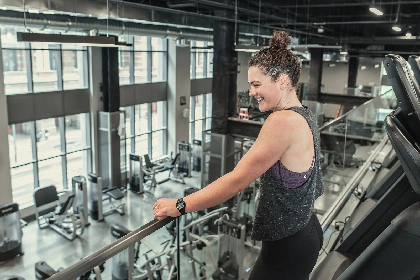 A woman smiling on a treadmill in a modern gym with large windows and various exercise equipment.