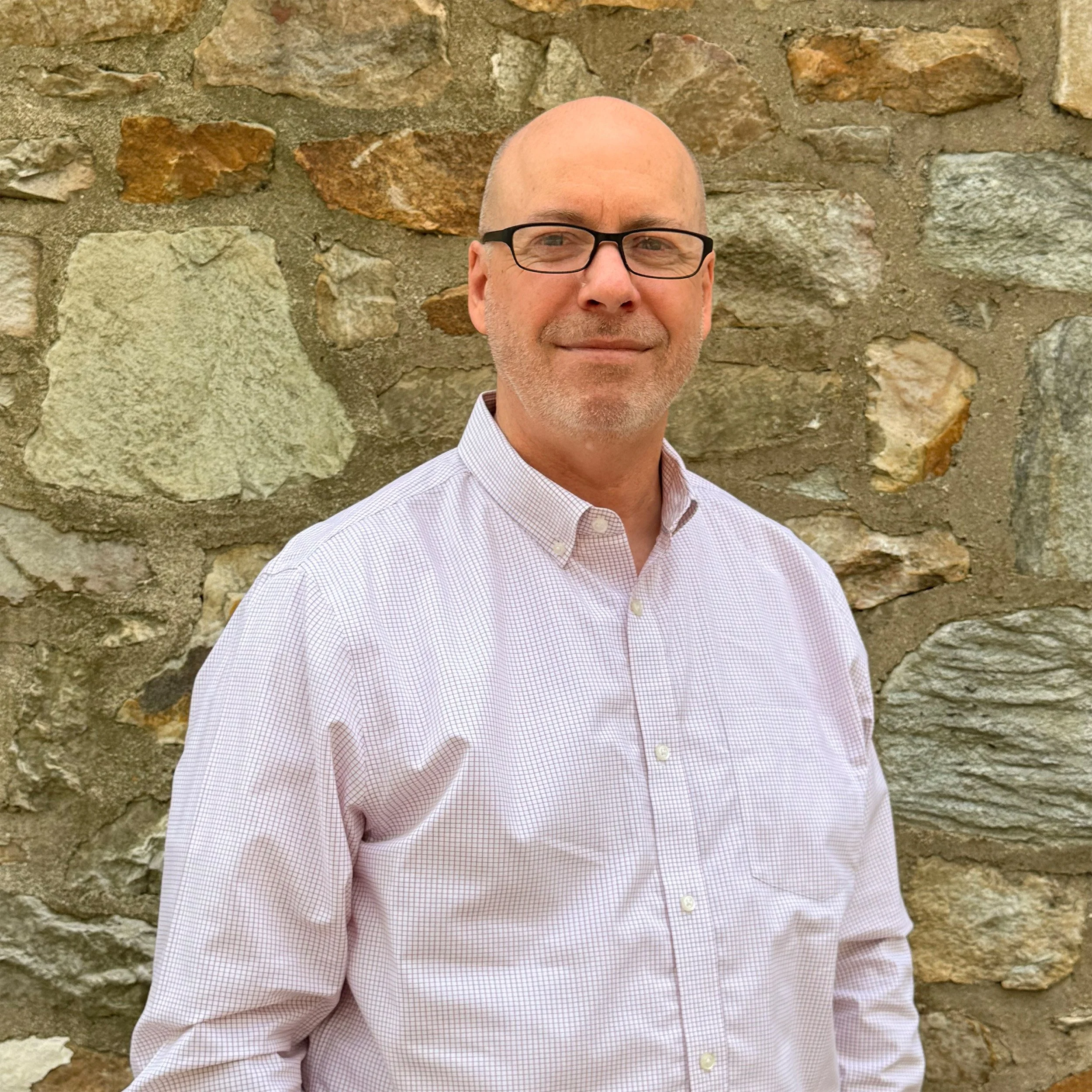 A middle-aged man with glasses and a light checkered shirt in front of a stone wall.