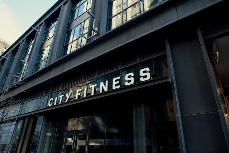 Exterior of a modern building with large windows, displaying a sign that reads 'CITY FITNESS' in white letters.