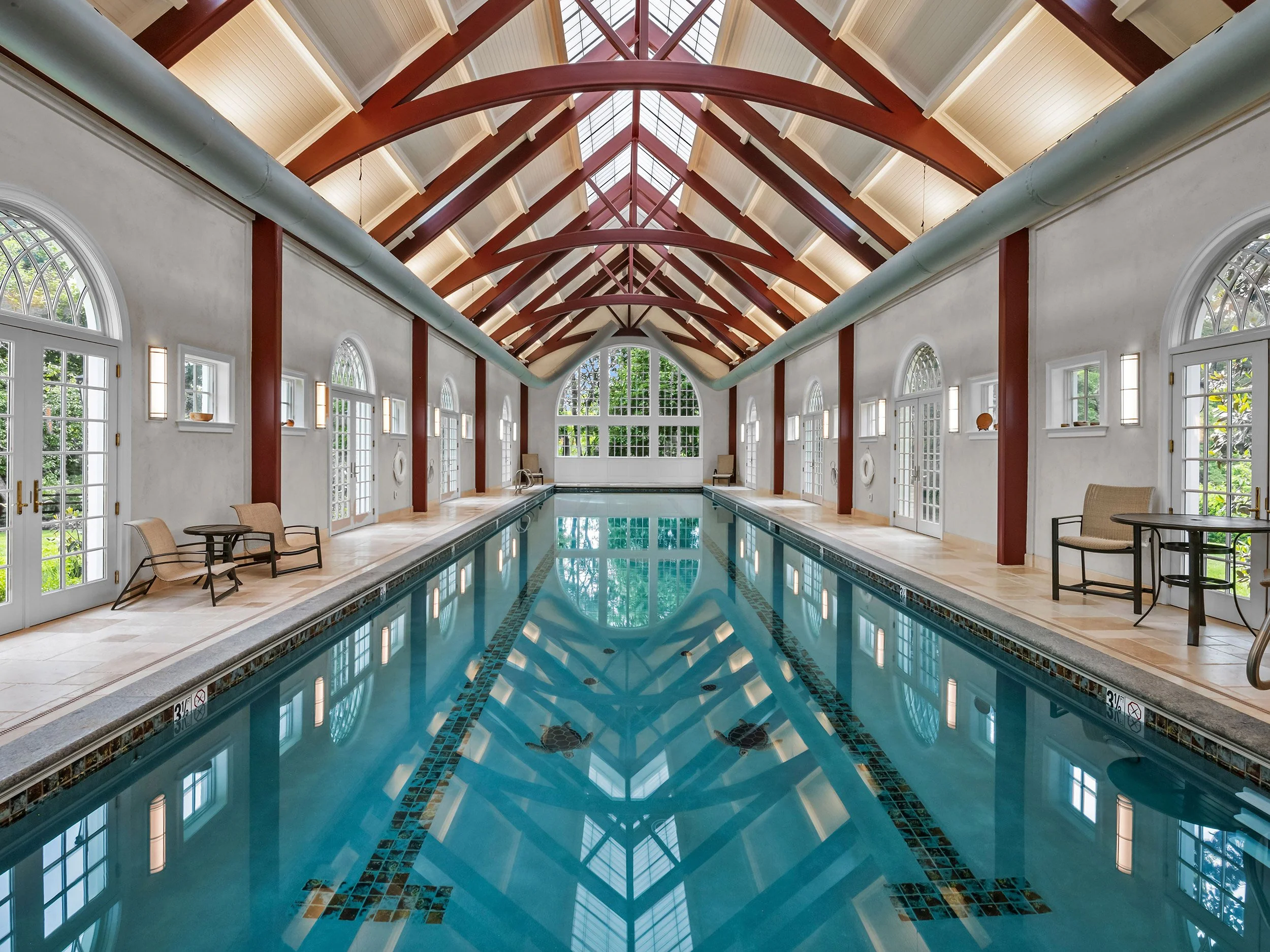 Indoor swimming pool area with large windows, high vaulted ceiling with exposed beams, and lounge chairs along the sides.