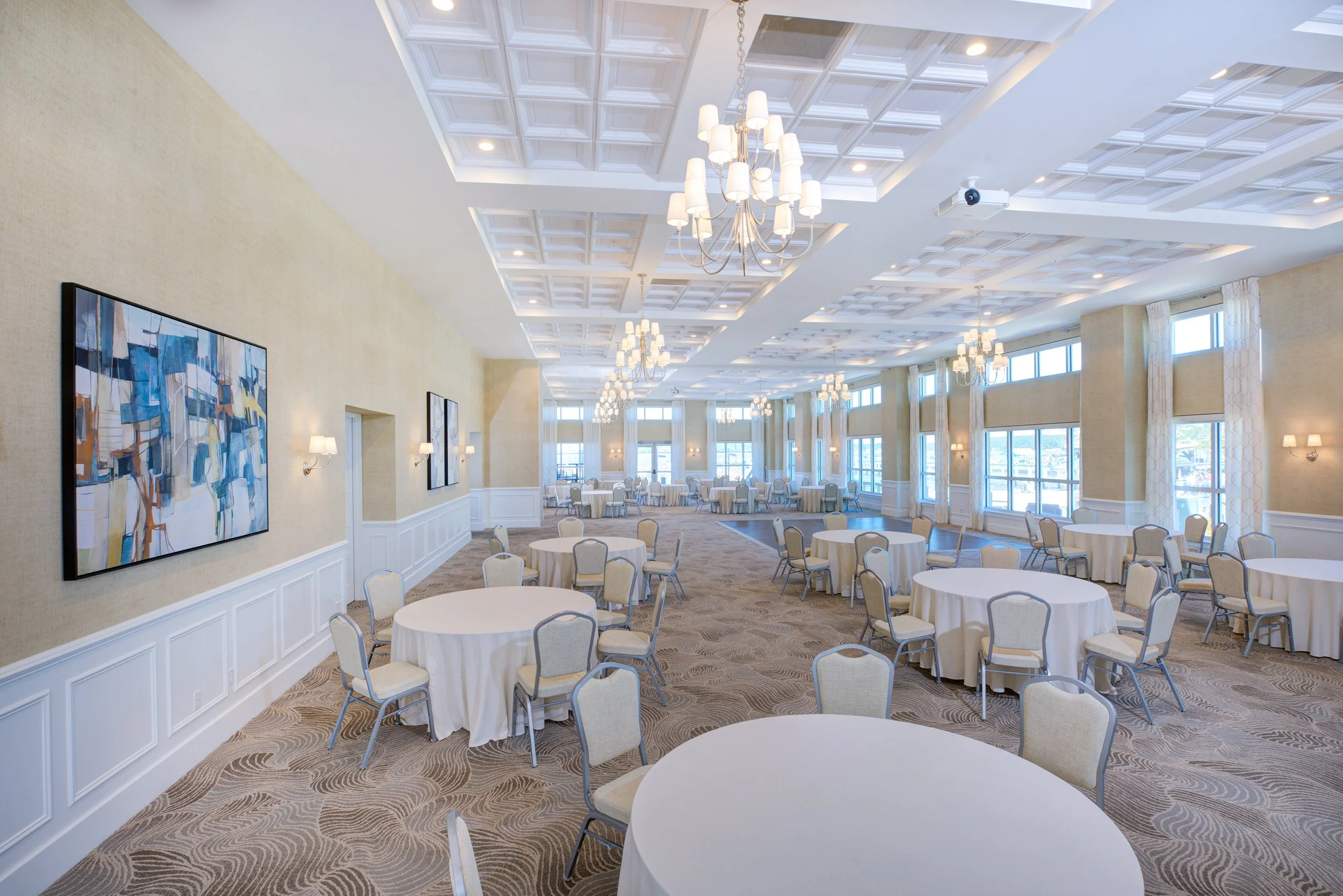Elegant banquet hall with round tables covered in white tablecloths, beige chairs, large windows with white curtains, abstract paintings on the walls, high ceiling with decorative panels, chandeliers, and ample natural light.