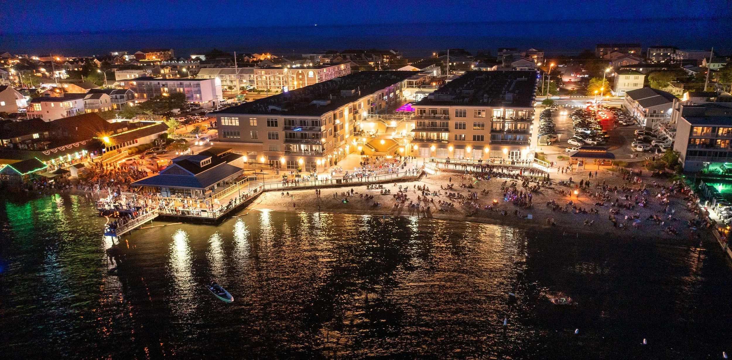 Nighttime aerial view of a lively beachside area with illuminated buildings, crowded sandy beach, and water with boats in the foreground.