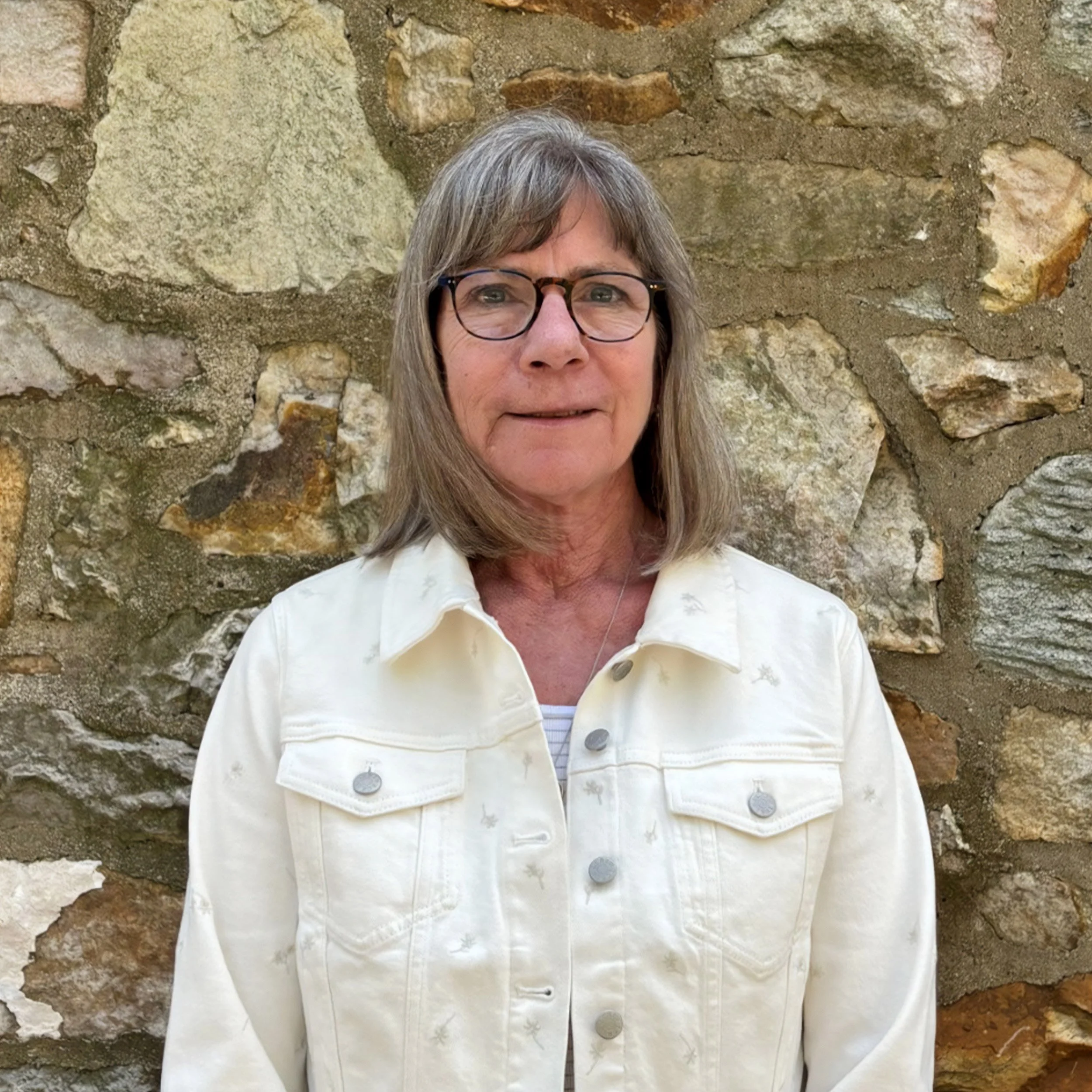 A woman with gray hair and glasses standing in front of a stone wall.