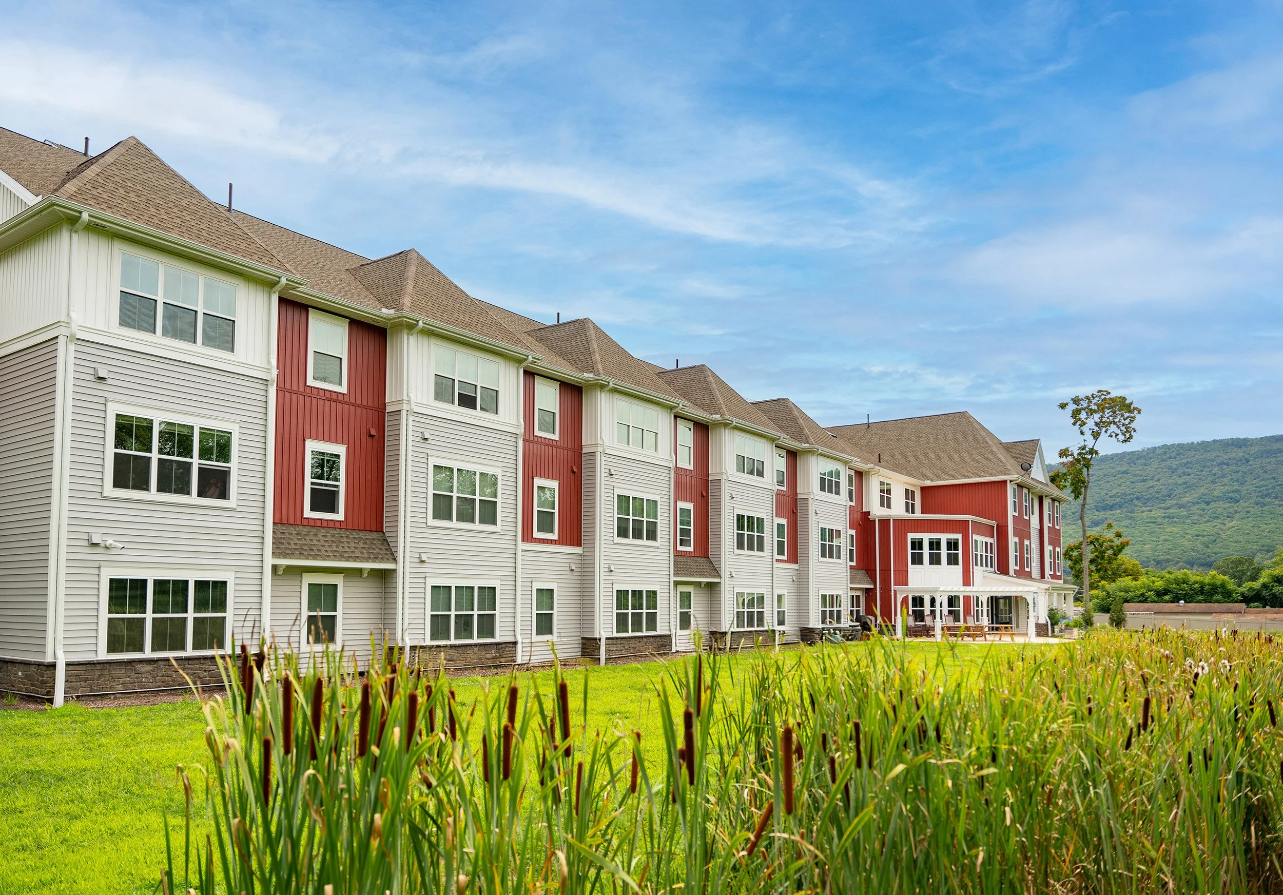 A multi-story residential building with beige and red siding, multiple windows, and a landscaped yard with green grass and tall plants, set against a backdrop of hills and a partly cloudy sky.