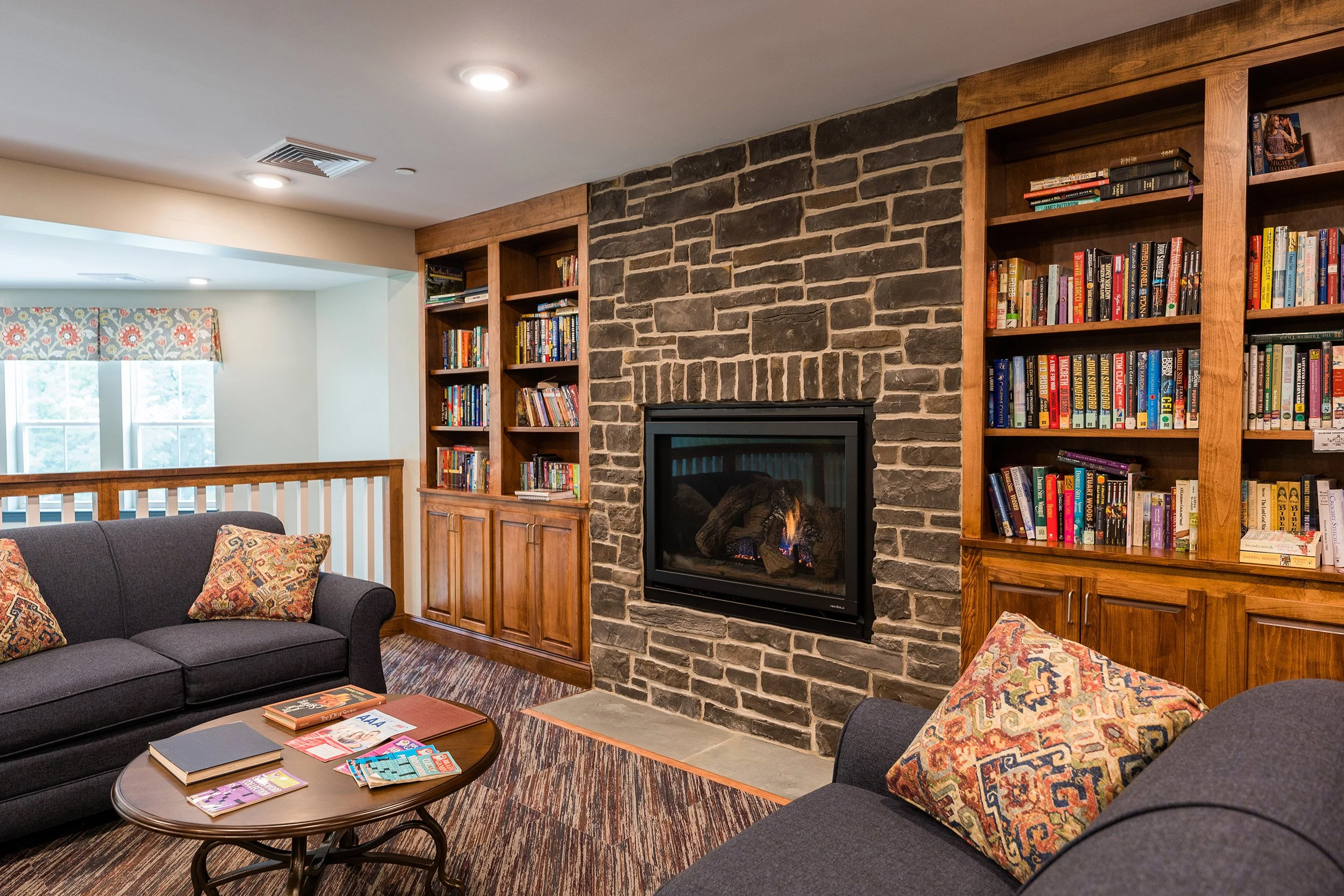 Lounge area with a brick fireplace, built-in wooden bookshelves filled with books, gray couches with patterned pillows, and a wooden coffee table with magazines and books.