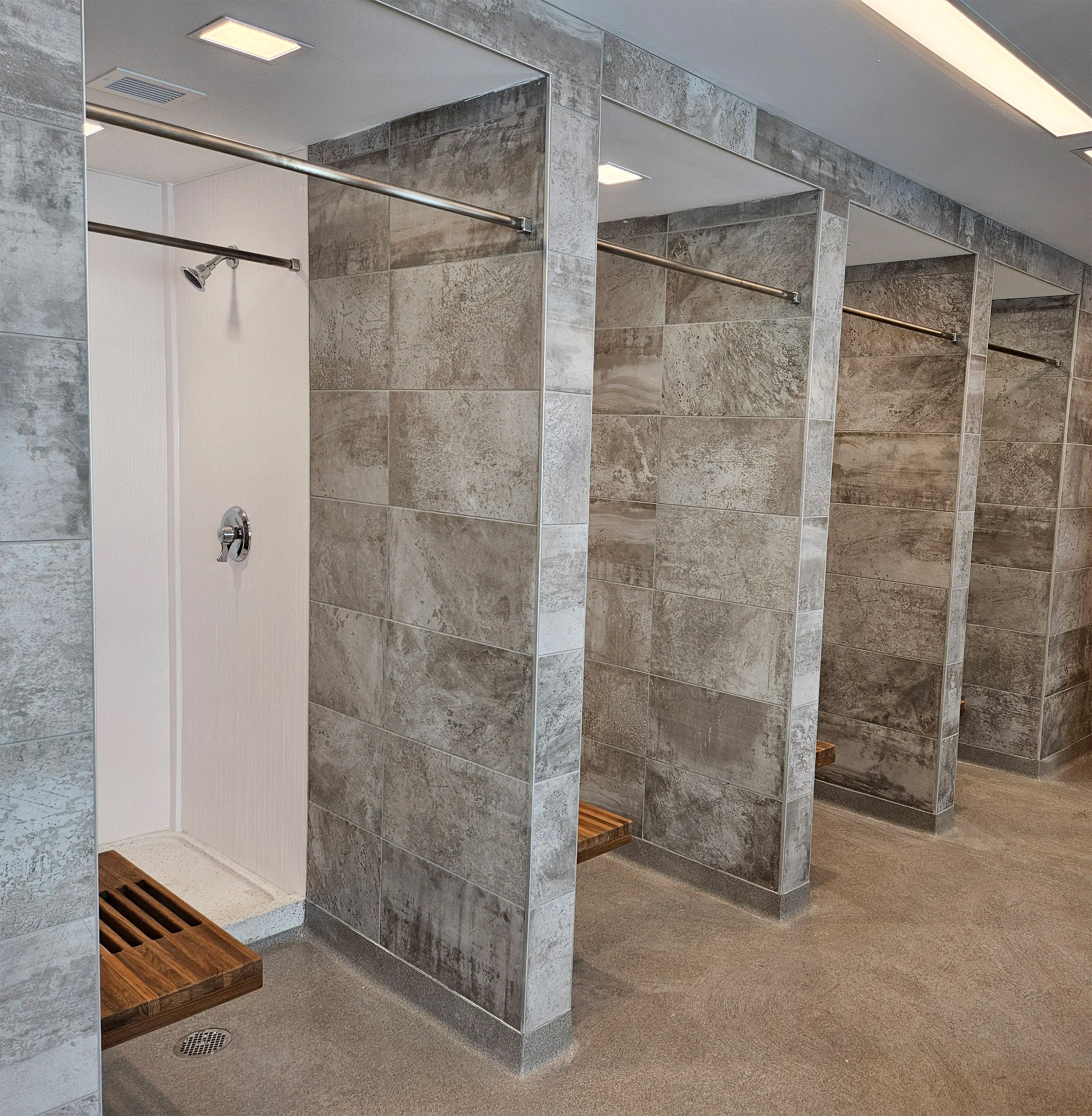 Dormitory shower space with three individual showers separated by tiled walls, each with a wooden slatted bench, a showerhead, and a drain on the floor.