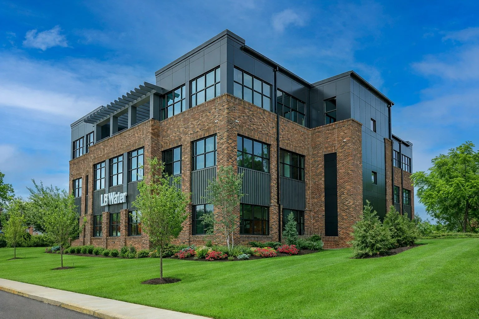 Modern brick and black metal building with large windows, surrounded by trees, grass, and flowers, under a blue sky.