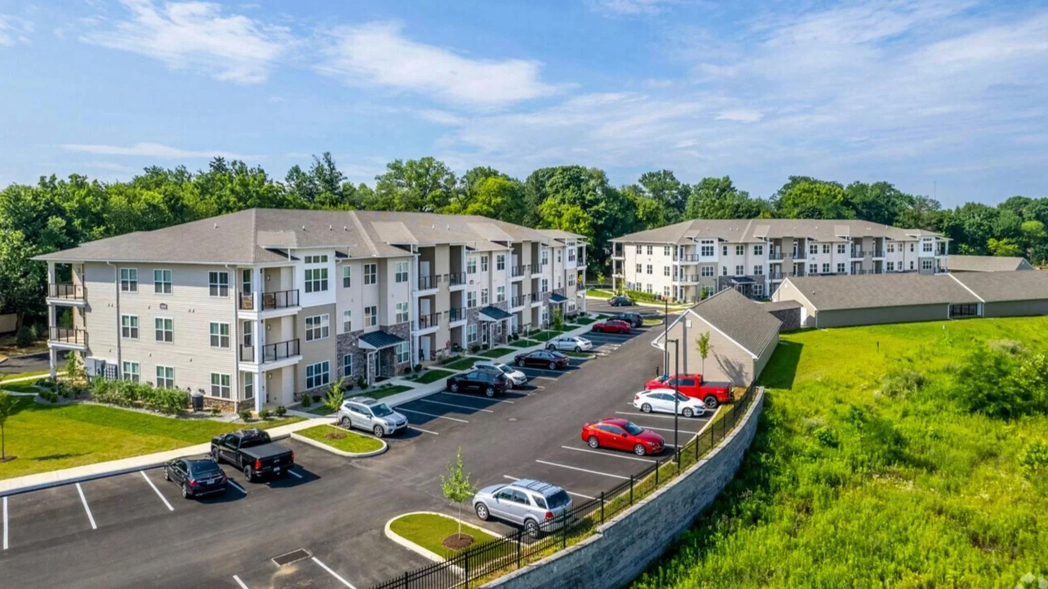 Aerial view of a modern apartment complex with four-story buildings, parking lot with cars, surrounded by green trees and a grassy area under a partly cloudy sky.
