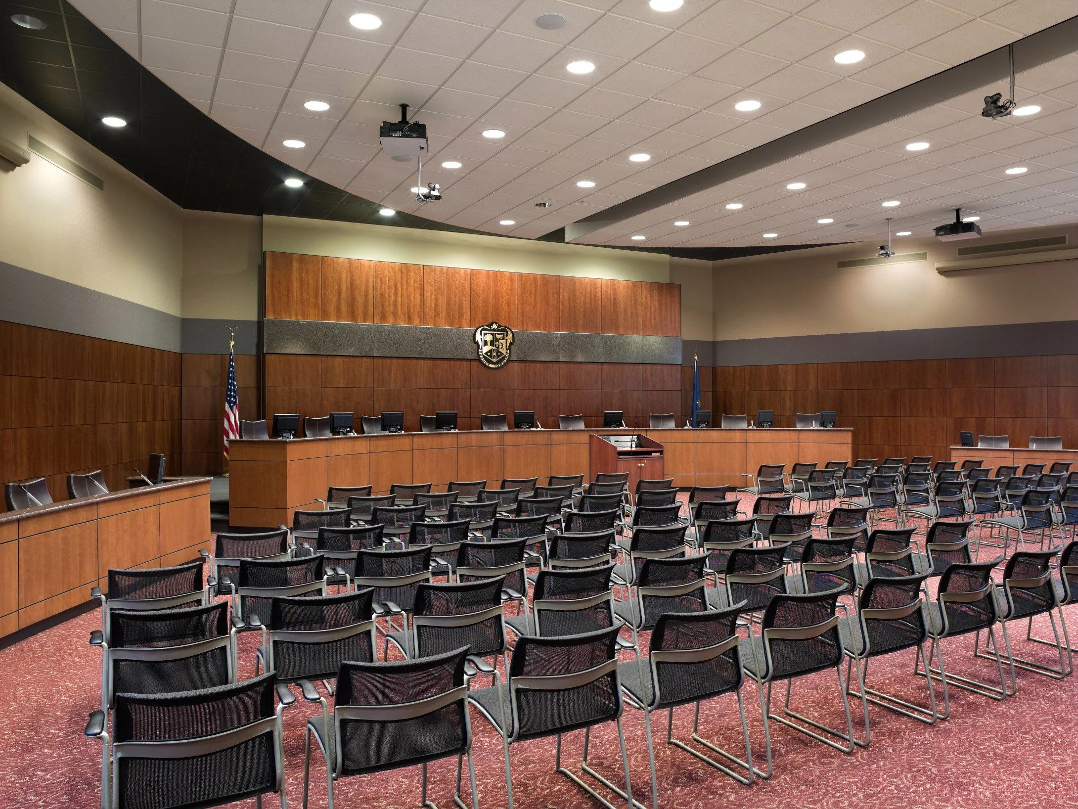 Empty meeting room with multiple chairs arranged facing a counter with monitors and flags.