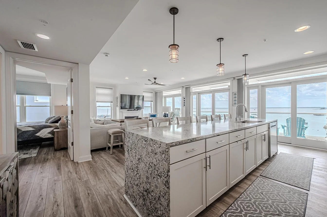 Open-concept living room and kitchen with large windows showing an ocean view. The kitchen has a granite island, white cabinets, and pendant lighting. The living area includes a TV and seating, with a bedroom visible through an open door.
