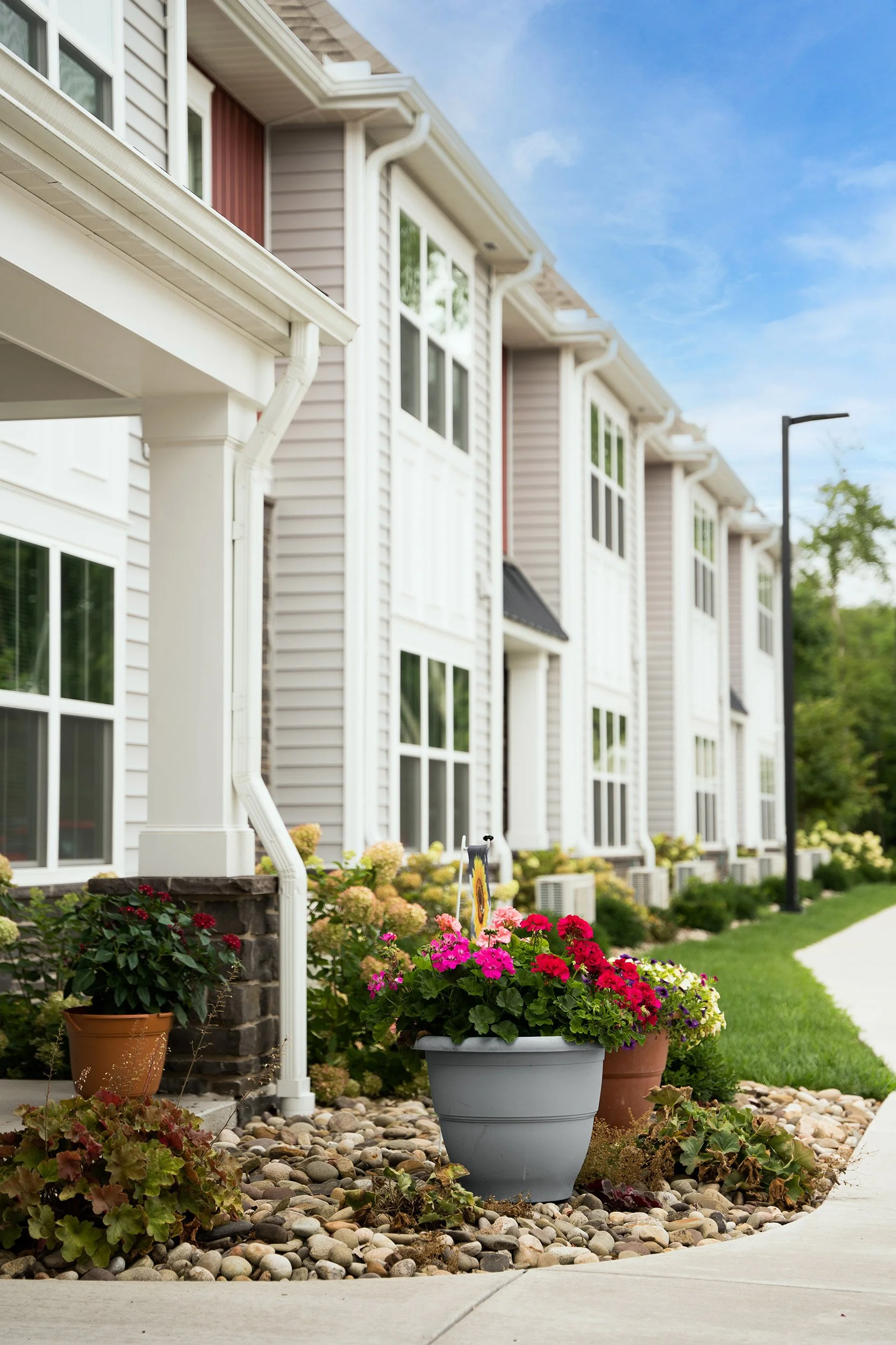 Residential building with flower beds and potted plants along the sidewalk, under a blue sky.