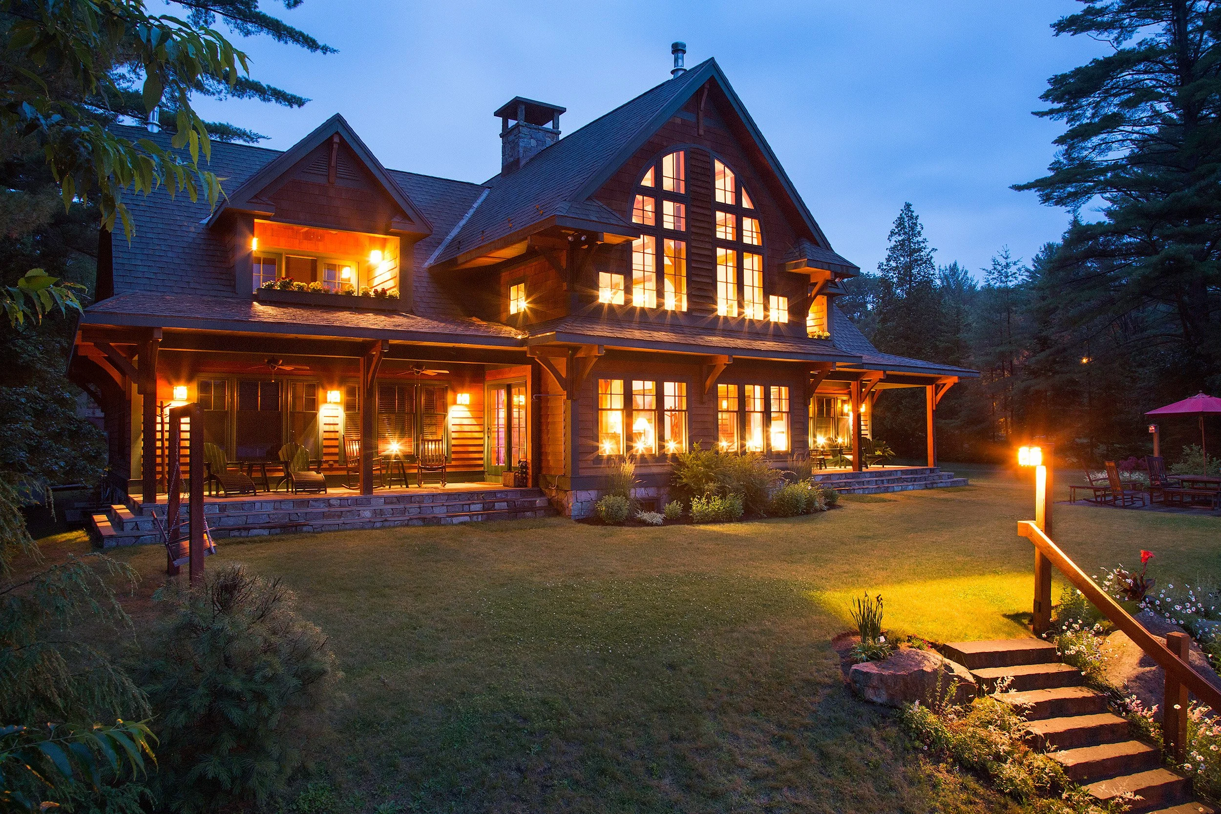 A large, multi-story Adirondack style house with warm lighting inside, situated in a wooded area during dusk. The house has a wooden exterior, multiple windows, a porch with seating, and a landscaped front yard with steps leading up to it.