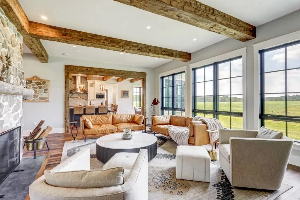 Spacious living room with leather sofas, armchairs, large windows with a scenic view, stone fireplace, and wooden beams on the ceiling.