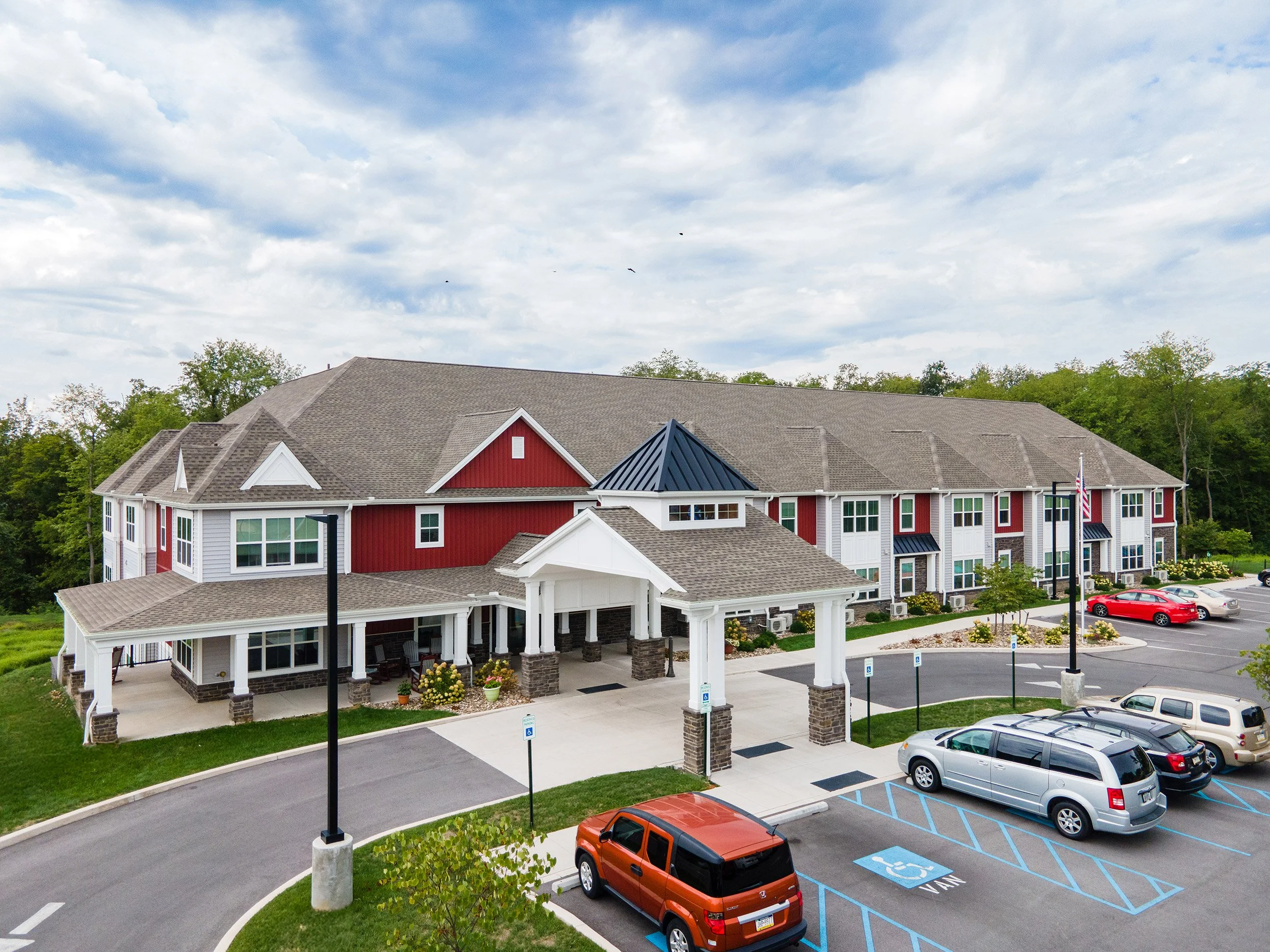 A large building with a combination of red and white exterior walls, surrounded by a parking lot with several cars, including a wheelchair accessible parking spot. The building has a covered entrance supported by stone pillars and is set against gree