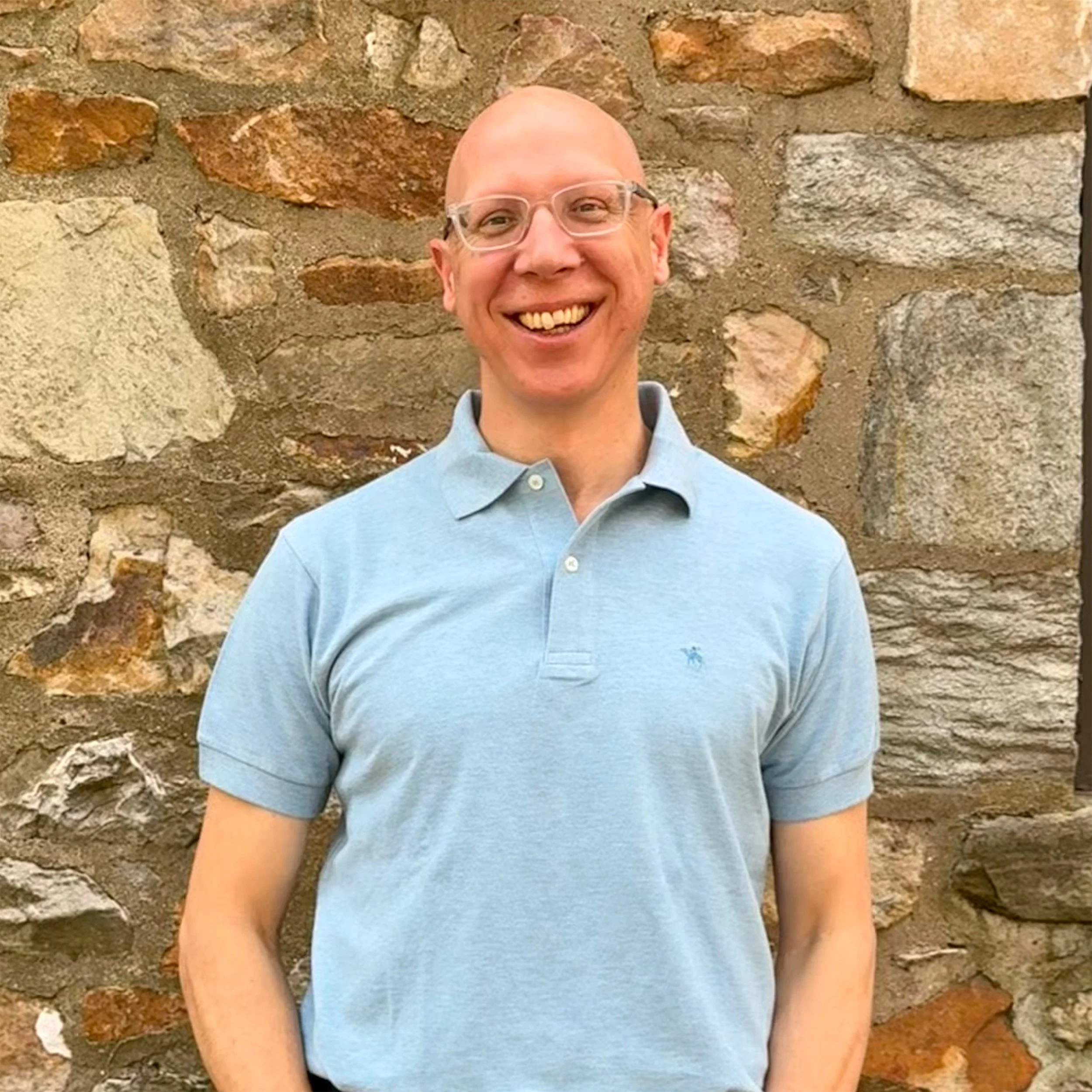 A man with glasses and a bald head smiling, standing in front of a stone wall.