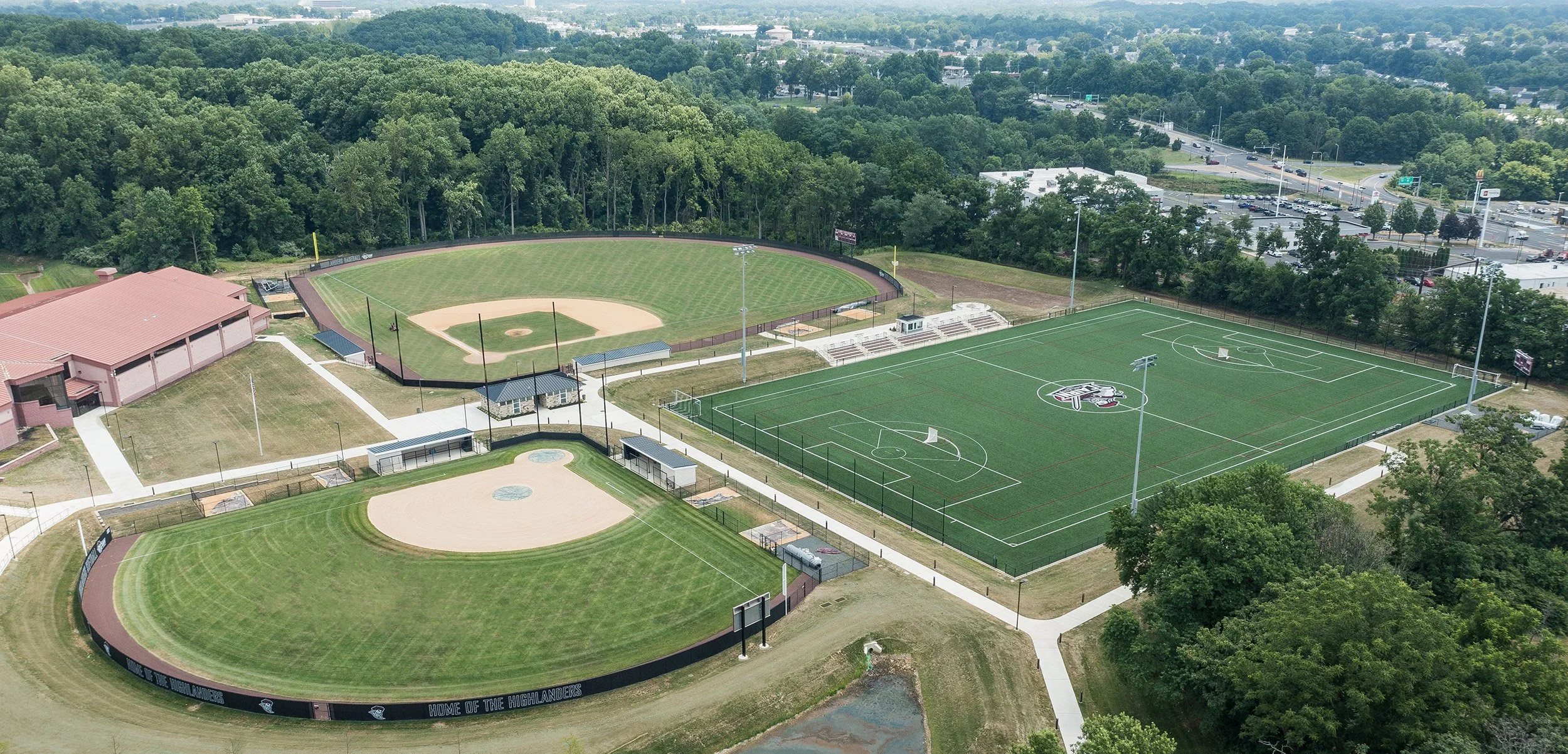 An aerial view of a sports complex featuring a baseball field, a soccer field, and a track with seating areas, surrounded by trees and parking lots.