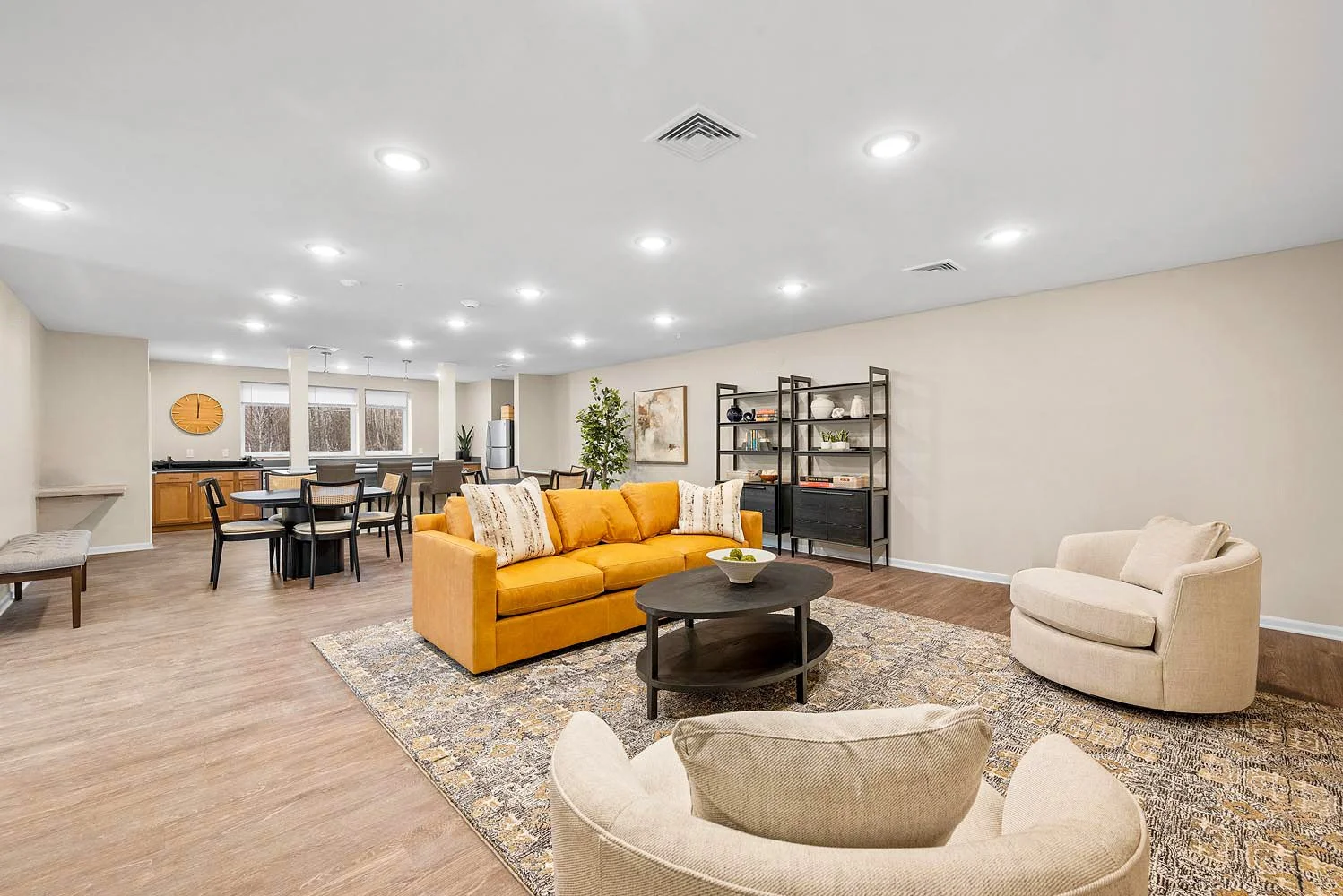 Open-concept living room and dining area with beige walls, wood flooring, a yellow sofa with patterned pillows, a beige armchair, a black coffee table with a bowl of fruit, a black bookshelf, and a rug; in the background, a kitchen with a breakfast b