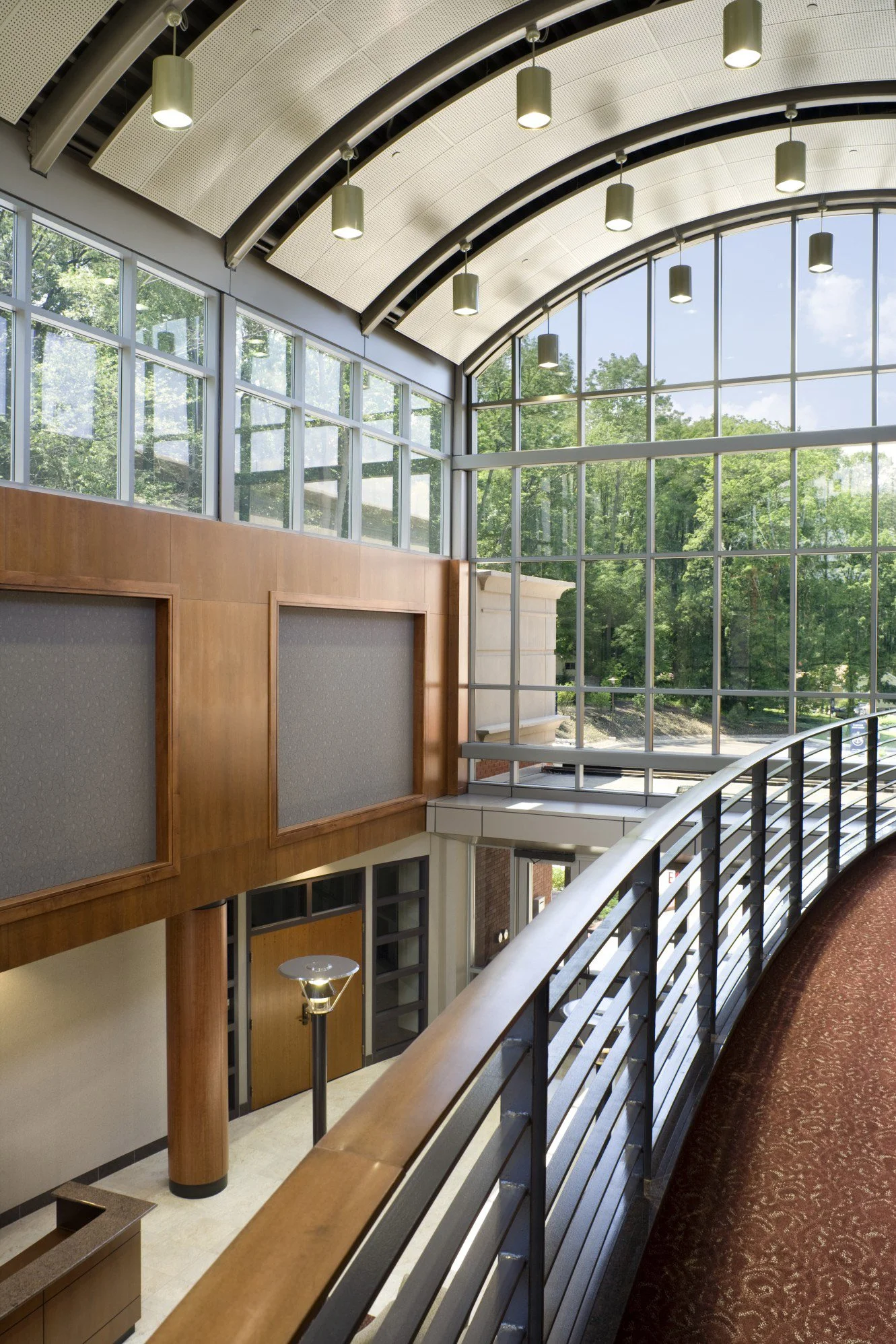 Interior view of a modern building with large glass windows, wooden panel walls, and a curved stair railing. Ceiling has hanging cylindrical light fixtures. Outside greenery is visible through the windows.