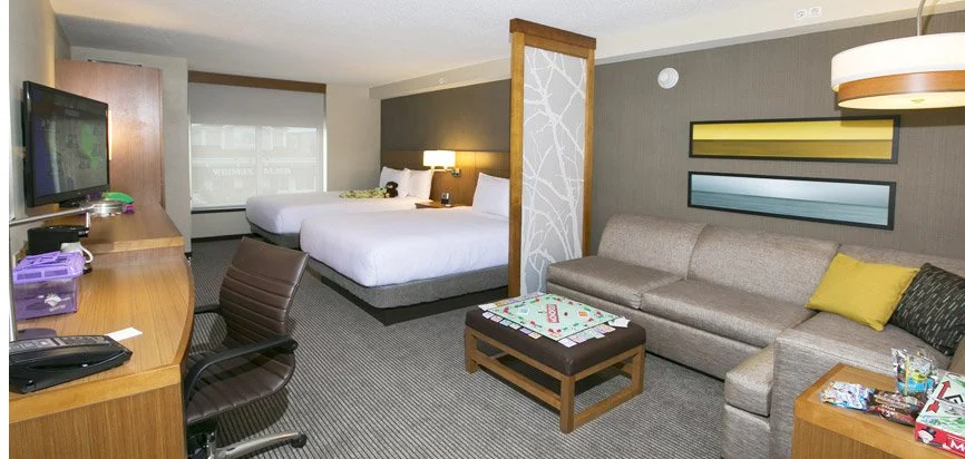 A hotel room with a sitting area and a bed. The sitting area has a beige couch with yellow and dark pillows, a wooden coffee table, and wall art. The bed has a headboard with a nightstand and lamp, and there is a desk with a chair and TV.