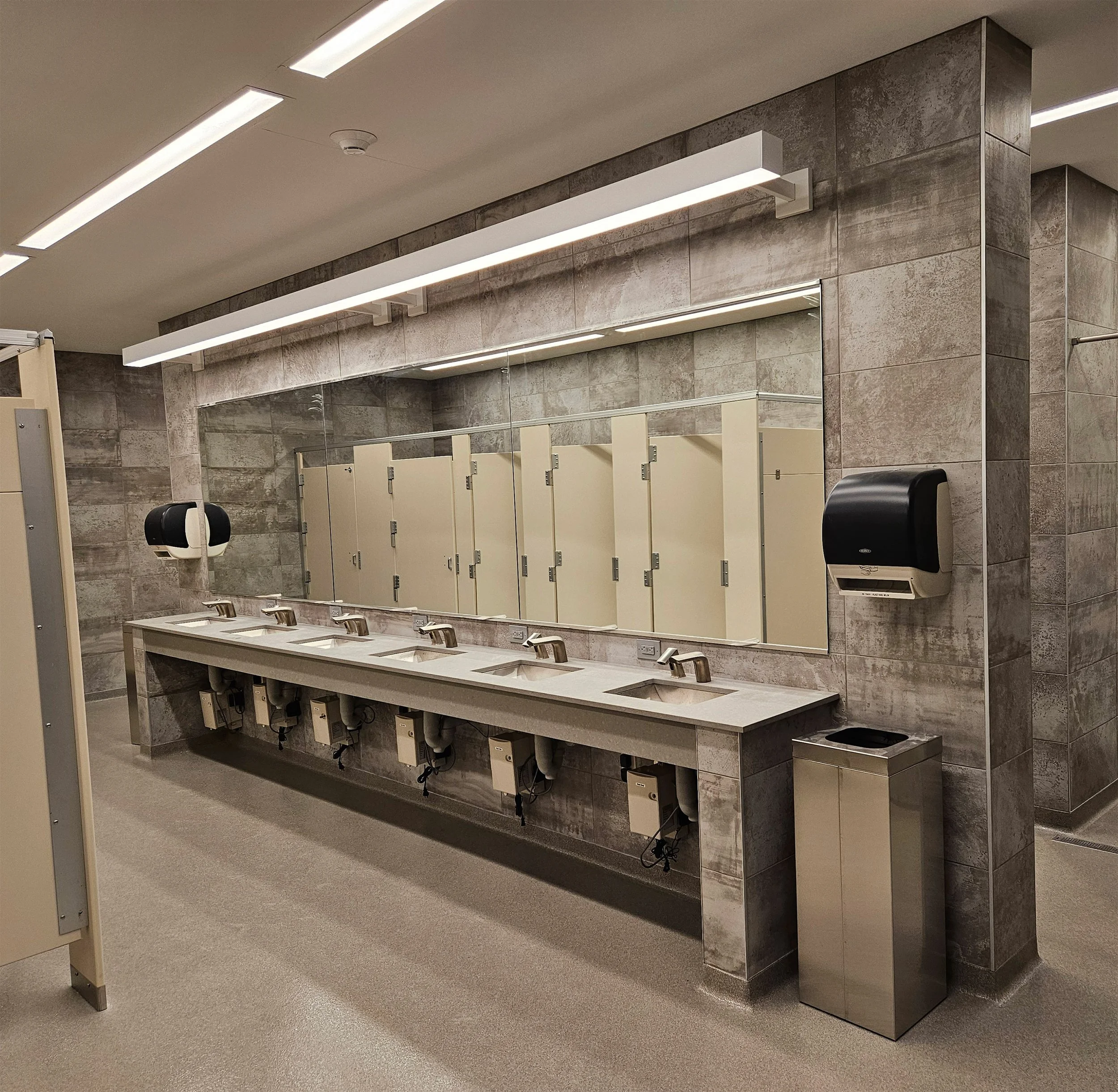 Dormitory bathroom with six sinks under a large mirror, soap dispensers on walls, paper towel dispenser, and trash bin, with beige stalls in the background.
