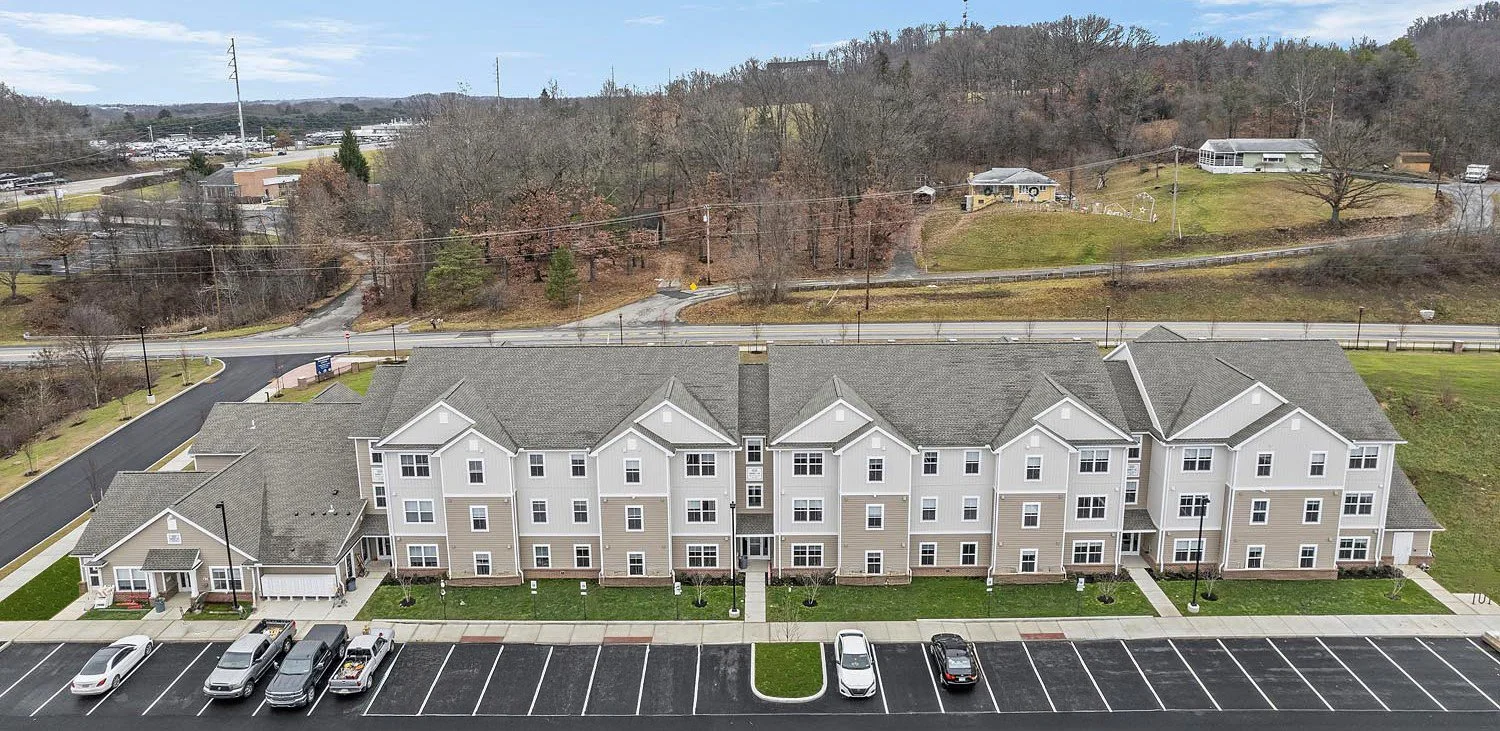 Aerial view of a residential apartment complex with parking lot, surrounded by trees and hills.