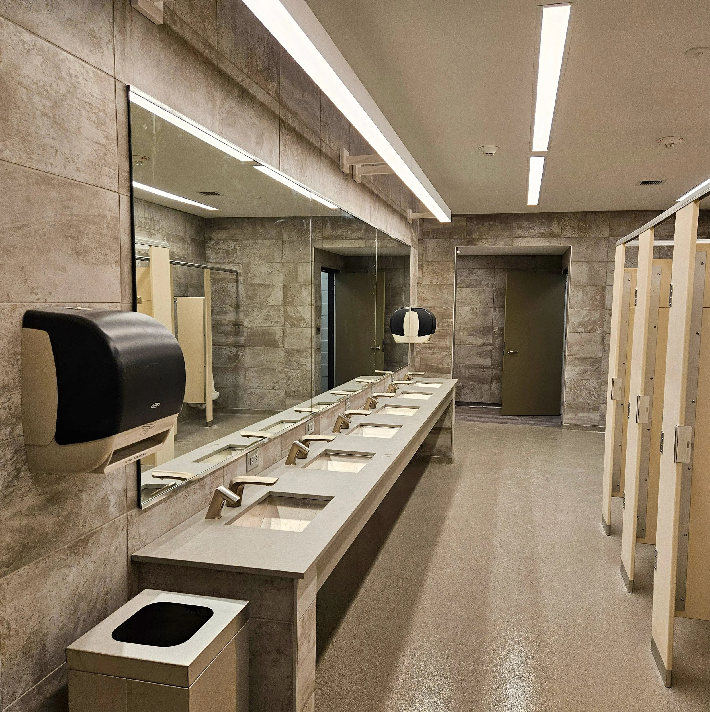 Dormitory bathroom with six sinks and a large mirror on a stone wall. There are two stalls with beige doors and a trash can near the sinks.