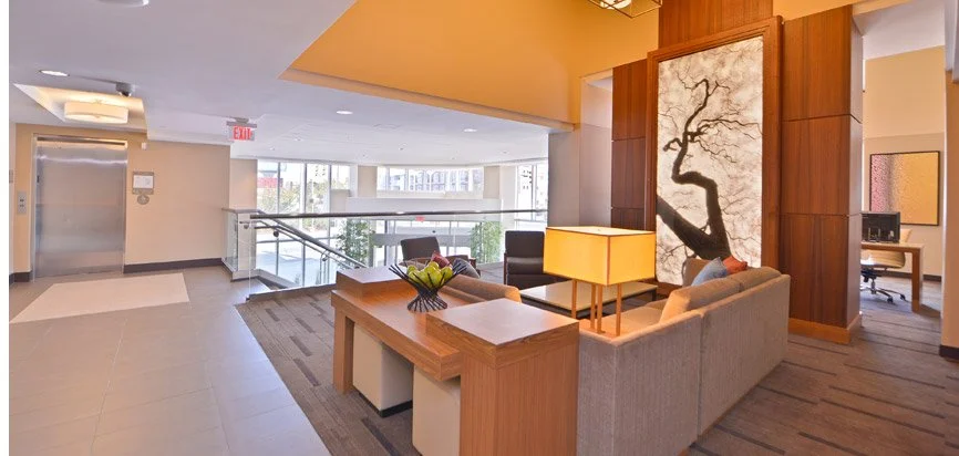 A hotel lobby area with seating, an elevator, large windows, and a decorative tree painting on the wall.