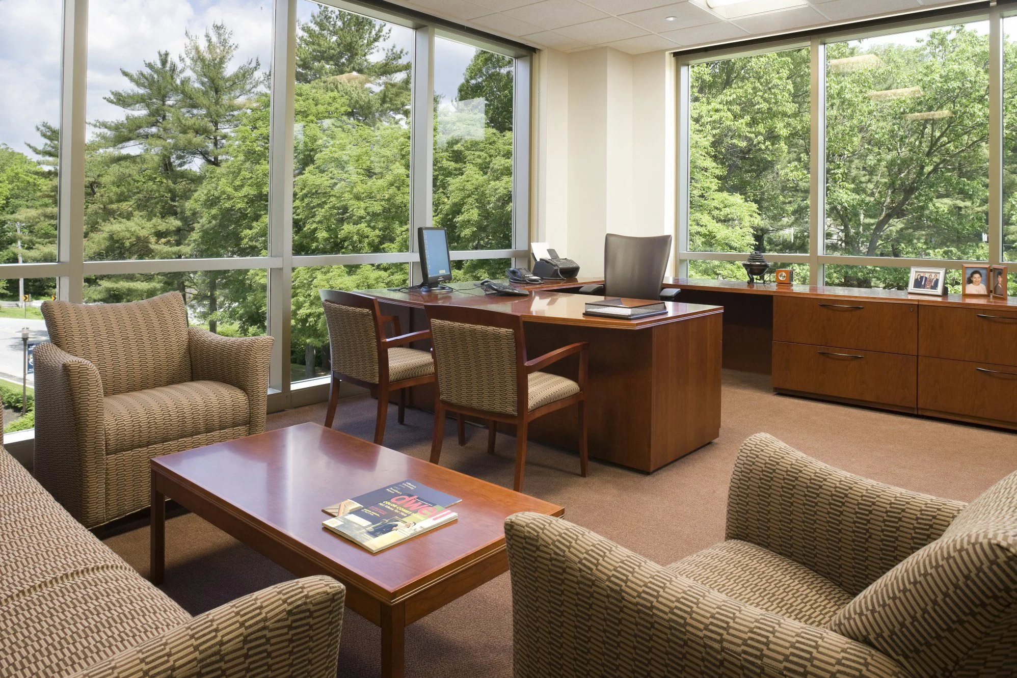 An office with a large wooden desk, a black office chair, two wooden chairs with patterned cushions, a sofa and armchair with matching patterned upholstery, a coffee table with magazines, a cabinet with framed photos, and large windows showing green 