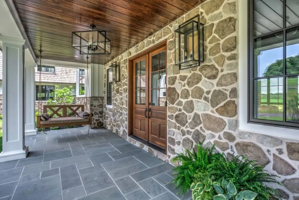Covered porch with stone wall exterior, wooden double doors, large windows, hanging lantern lights, a porch swing, and lush green plants.