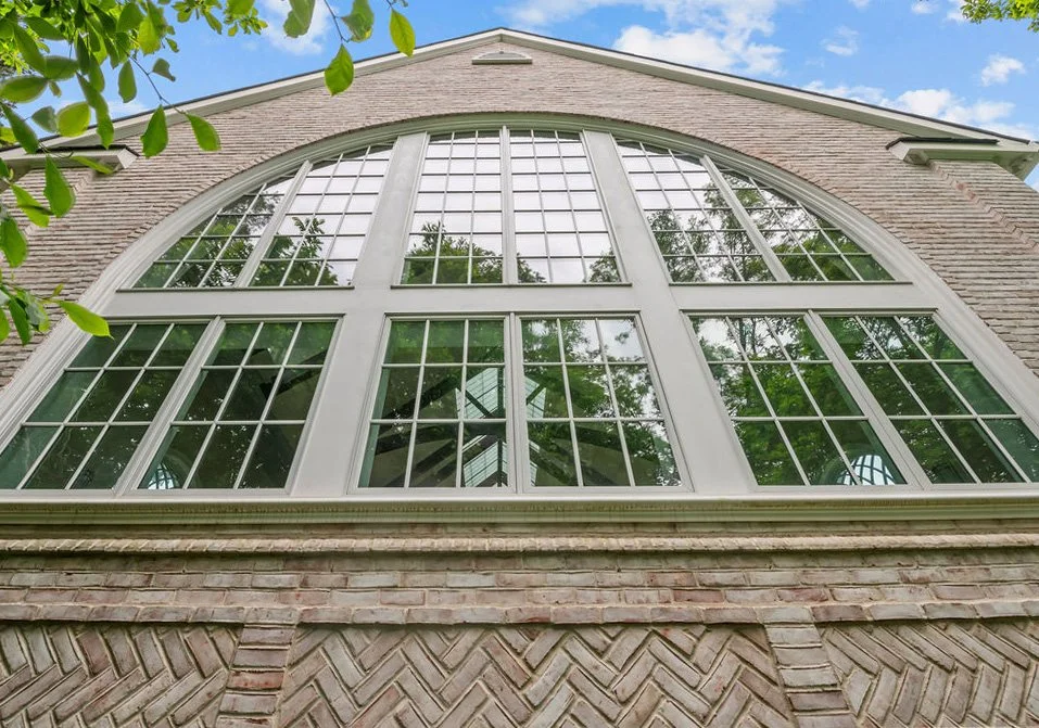 Large arched window with multiple panes on the brick exterior of a house, with trees and blue sky reflected in the glass.