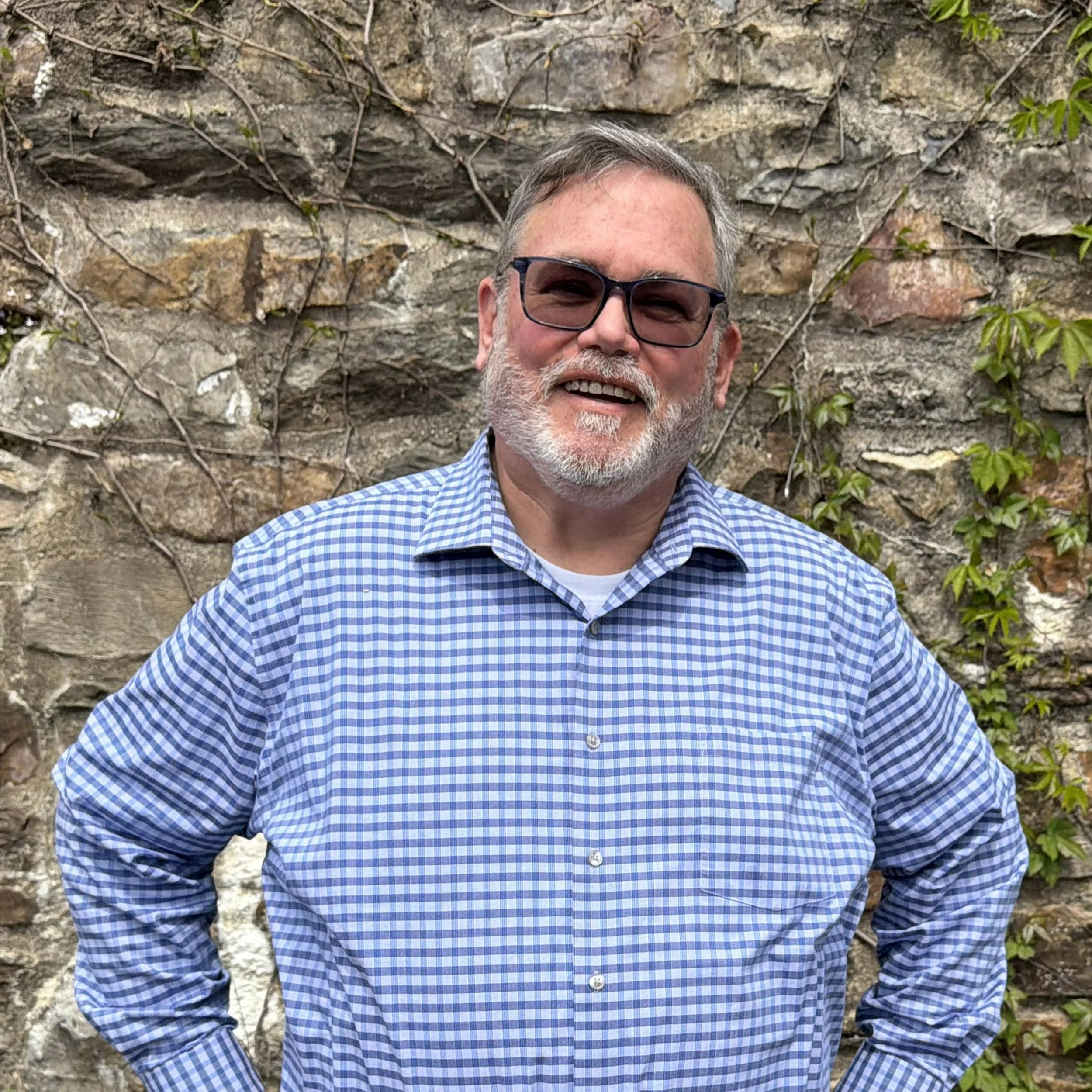 A smiling man with gray hair, beard, and glasses wearing a blue checkered shirt, standing outdoors in front of a stone wall with green ivy.