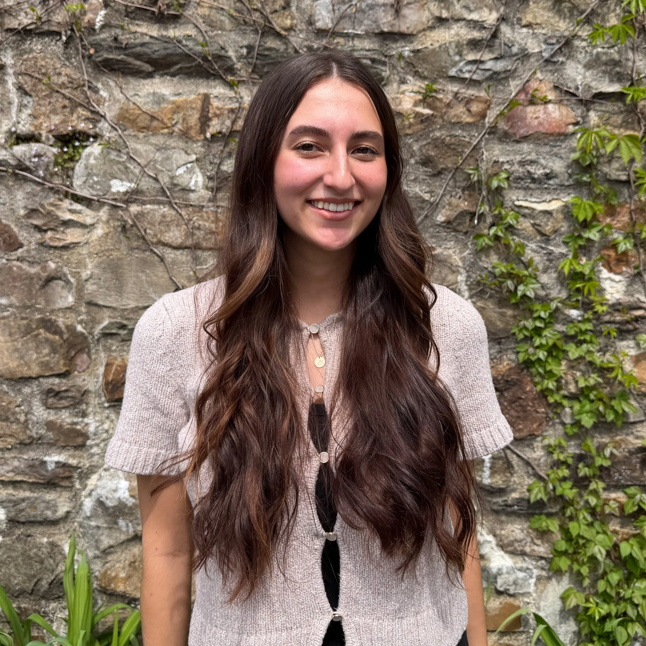 Close-up of a young woman with long brown hair, smiling, wearing a light beige cardigan and standing in front of a stone wall with green plants.