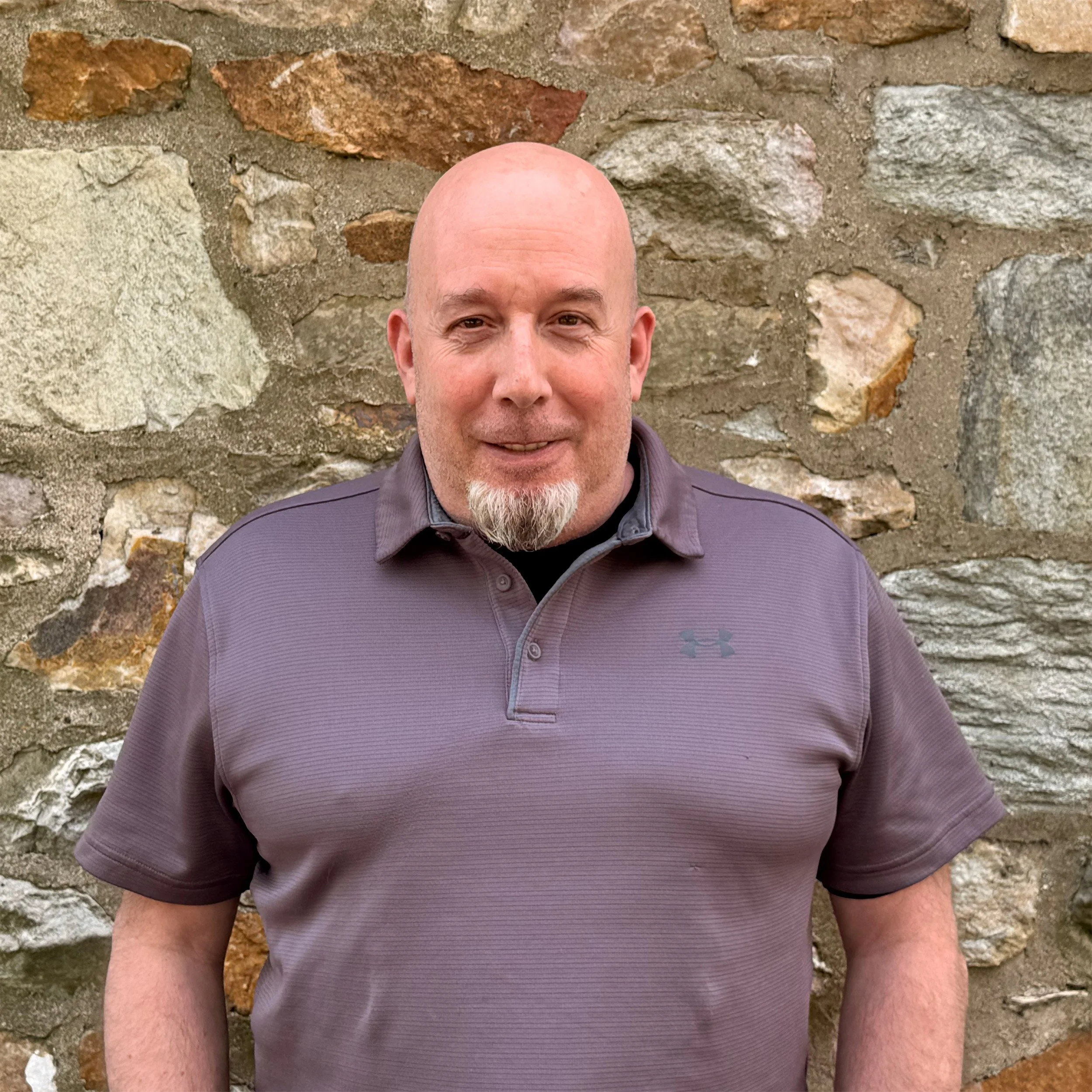 A close-up of a smiling man with a bald head and a goatee, wearing a purple shirt, standing in front of a stone wall made of various sizes and colors of stones.