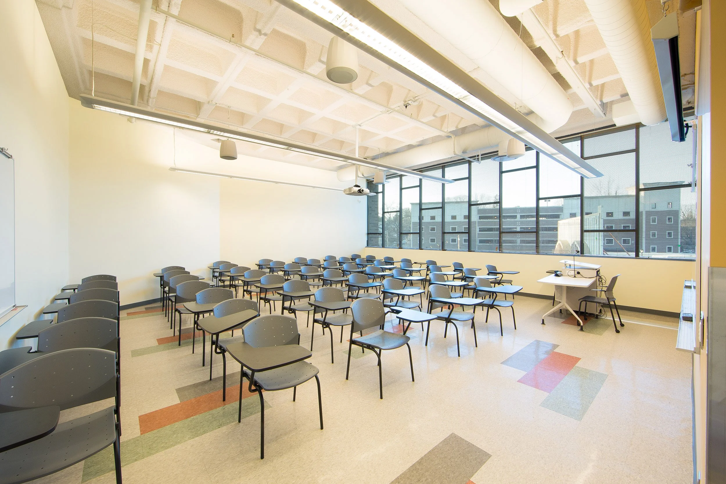 Empty college classroom with rows of gray chairs, a whiteboard, large windows, and a teacher's desk with a chair and projector at the front.