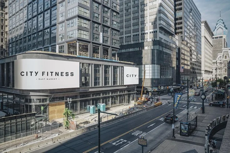 Modern city street with tall office buildings, a fitness center with large sign reading 'City Fitness', a few vehicles on the road, construction areas, and pedestrians on the sidewalk.